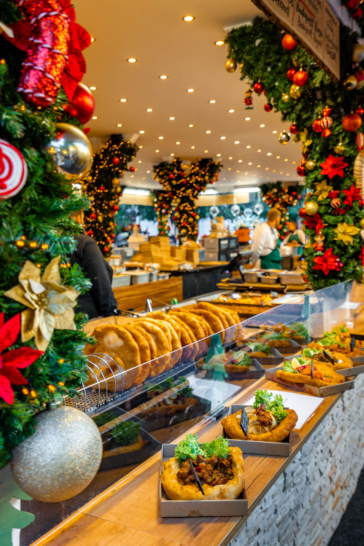 Street food counter at the Christmas market at Vörösmarty Square, Budapest, with local food displayed behind a glass case and festive garlands overhead.