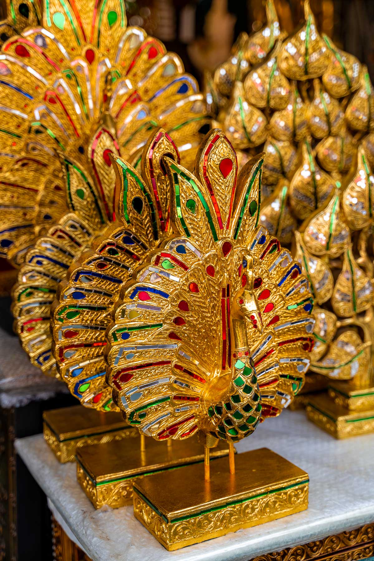 Golden peacock figurines with colorful red, green, and blue details displayed on a table at Chatuchak Weekend Market in Bangkok, stacked closely together in a souvenir stall.