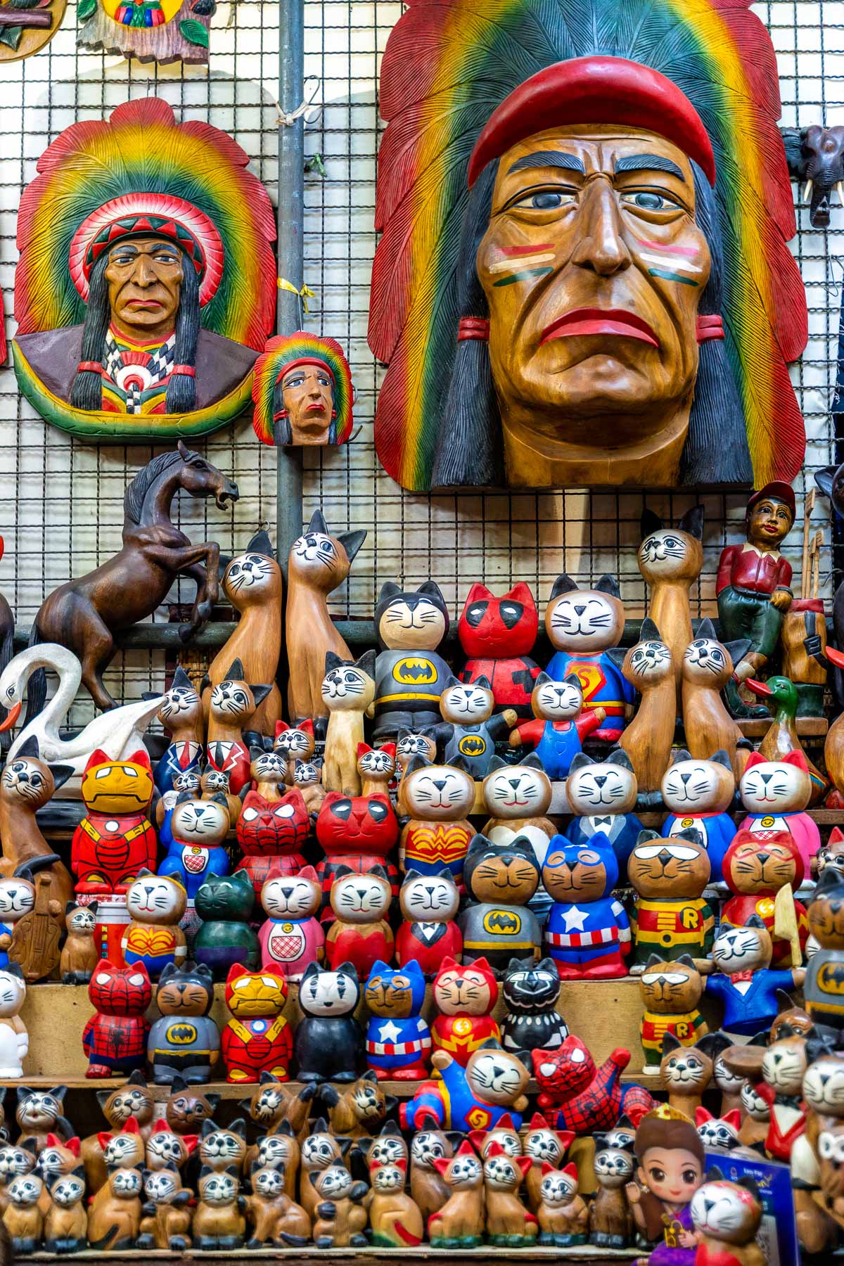 A souvenir stand at Chatuchak Weekend Market in Bangkok filled with wooden cat figurines dressed as superheroes, carved animal statues, and large decorative masks mounted on a grid wall behind them.