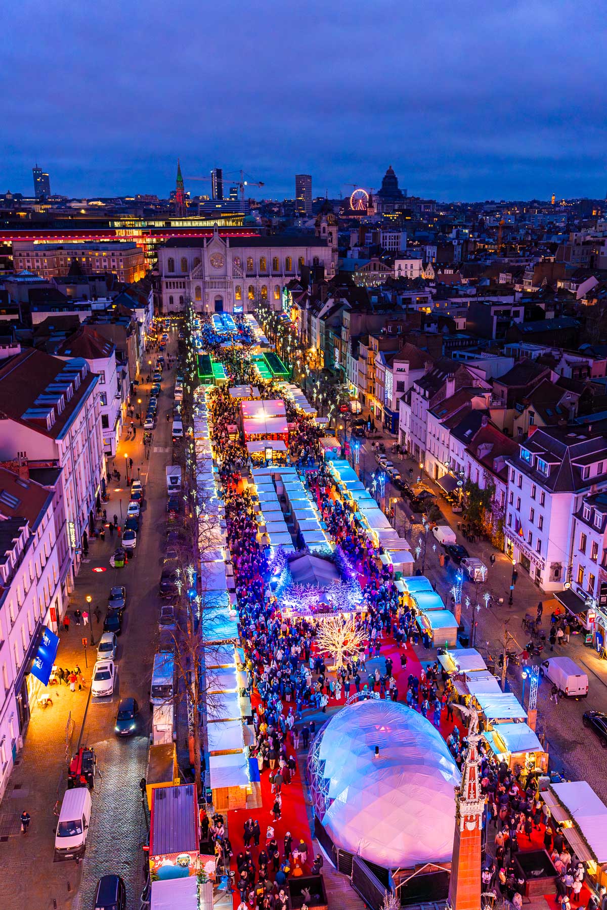 An aerial view of the Marché aux Poissons Christmas market in Brussels at dusk, with colorful lights, crowds, and a Ferris wheel in the distance.
