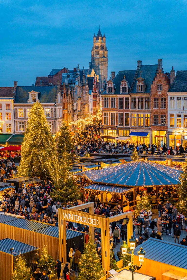 A busy evening at the Bruges Christmas market in Markt Square, with festive lights, Christmas trees, and crowds surrounded by historic Flemish buildings.