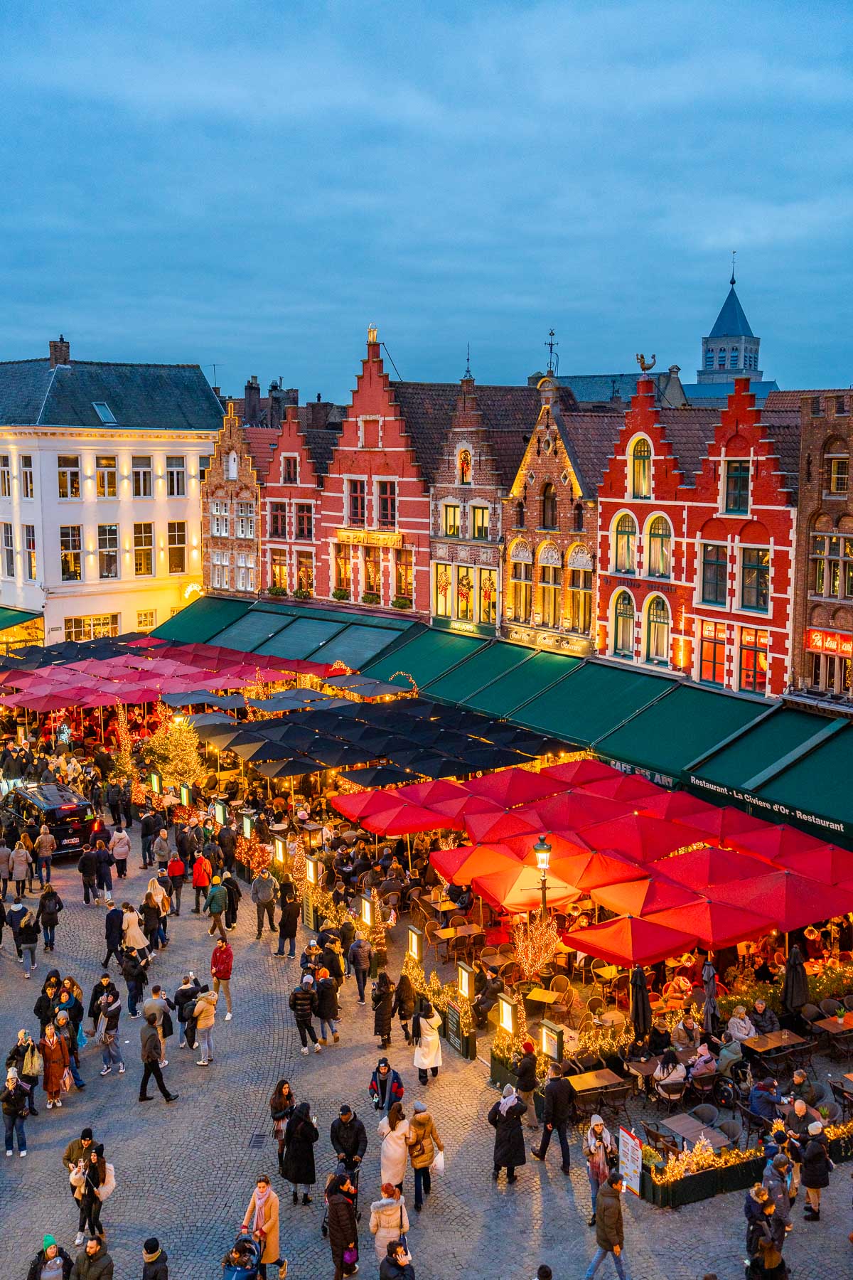 People walking around and dining under red and black umbrellas at the Bruges Christmas market in Markt Square, with colorful gabled houses in the background.
