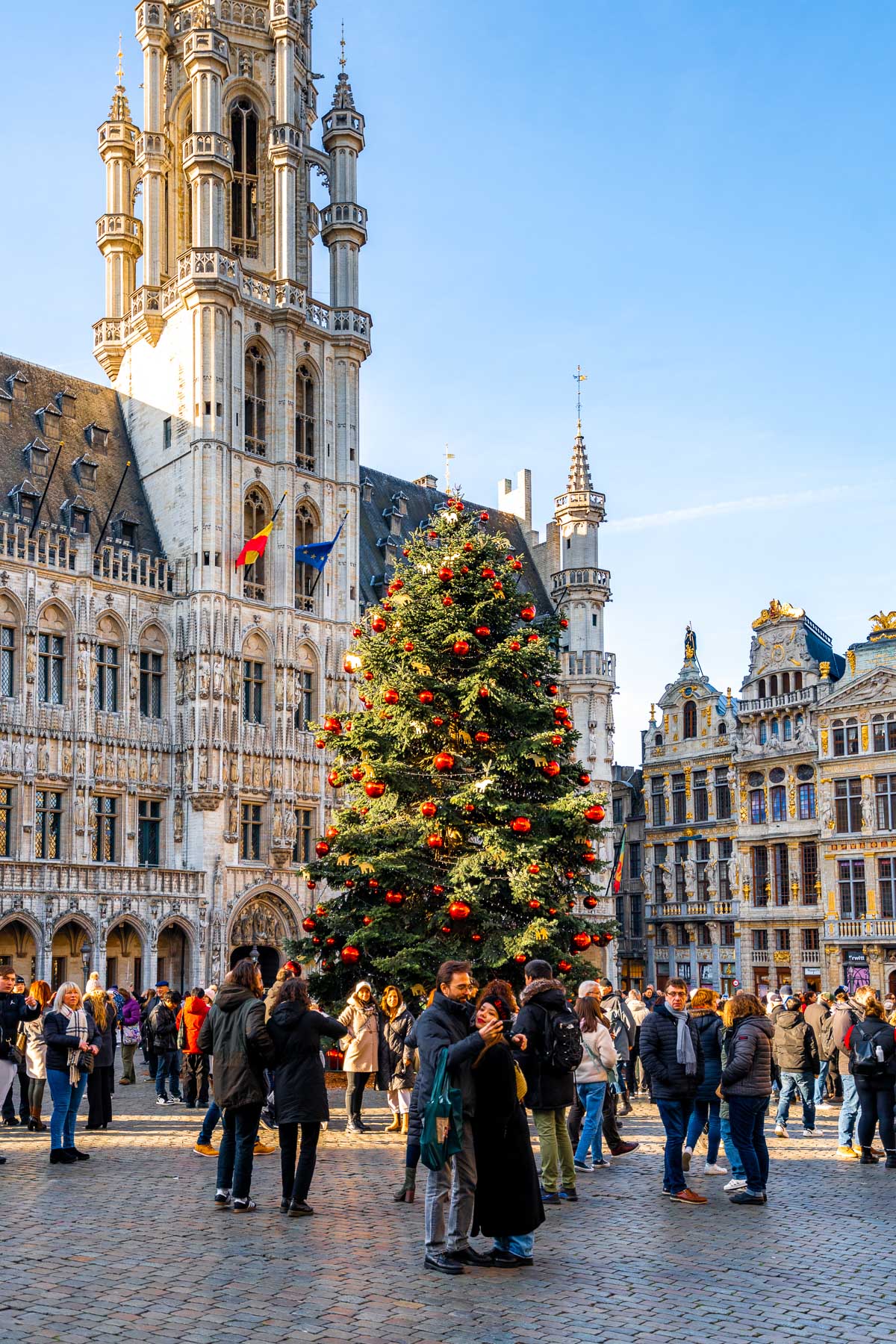 People gathered around the decorated Christmas tree at Grand Place in Brussels, surrounded by historic buildings and the Town Hall tower.