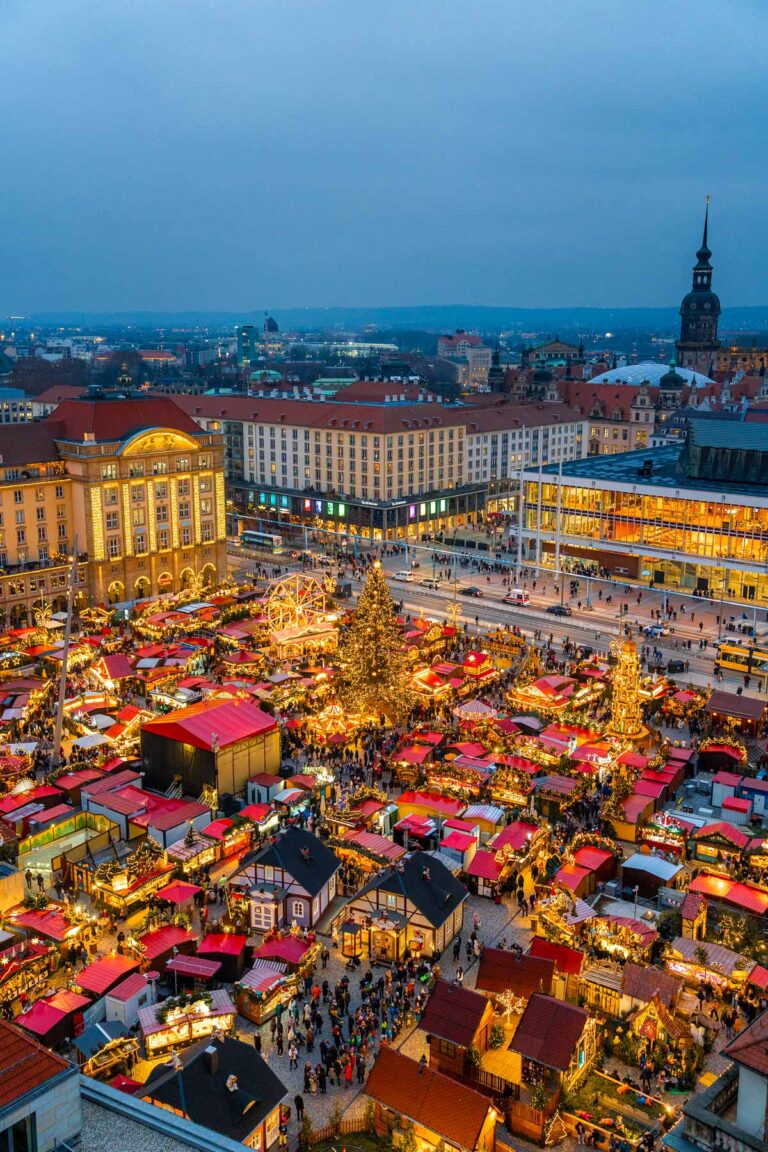 Aerial view of the Striezelmarkt in Dresden at dusk, filled with red-roofed stalls, a giant Christmas tree, and crowds enjoying the holiday atmosphere.