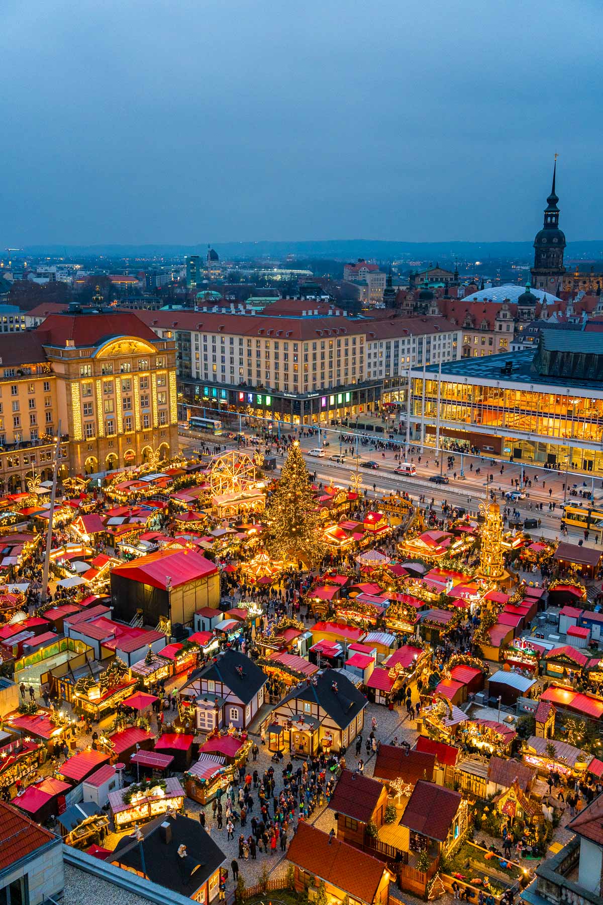 Aerial view of the Striezelmarkt in Dresden at dusk, filled with red-roofed stalls, a giant Christmas tree, and crowds enjoying the holiday atmosphere.