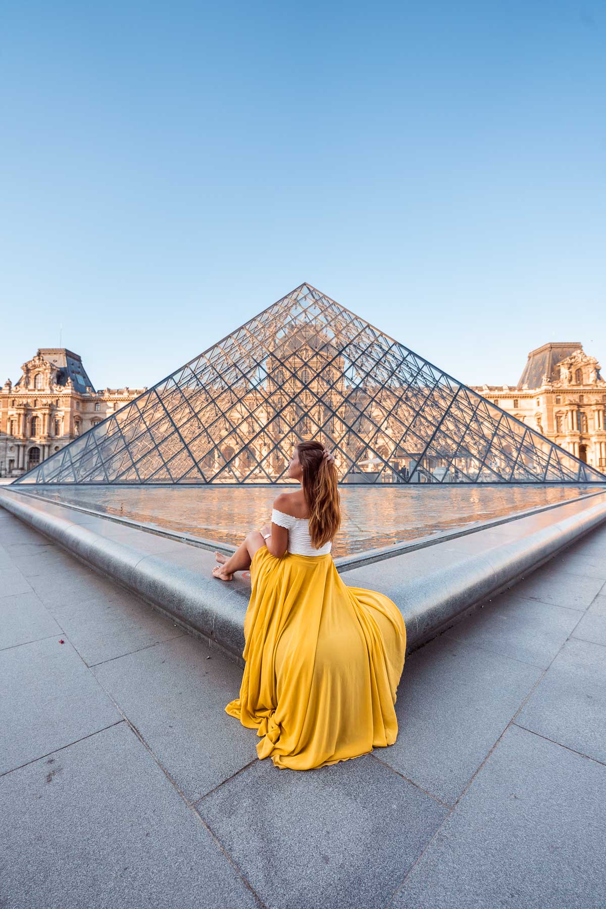 Girl in a yellow skirt sitting in front of one of the pyramids at the Louvre, one of the most Instagrammable places in Paris