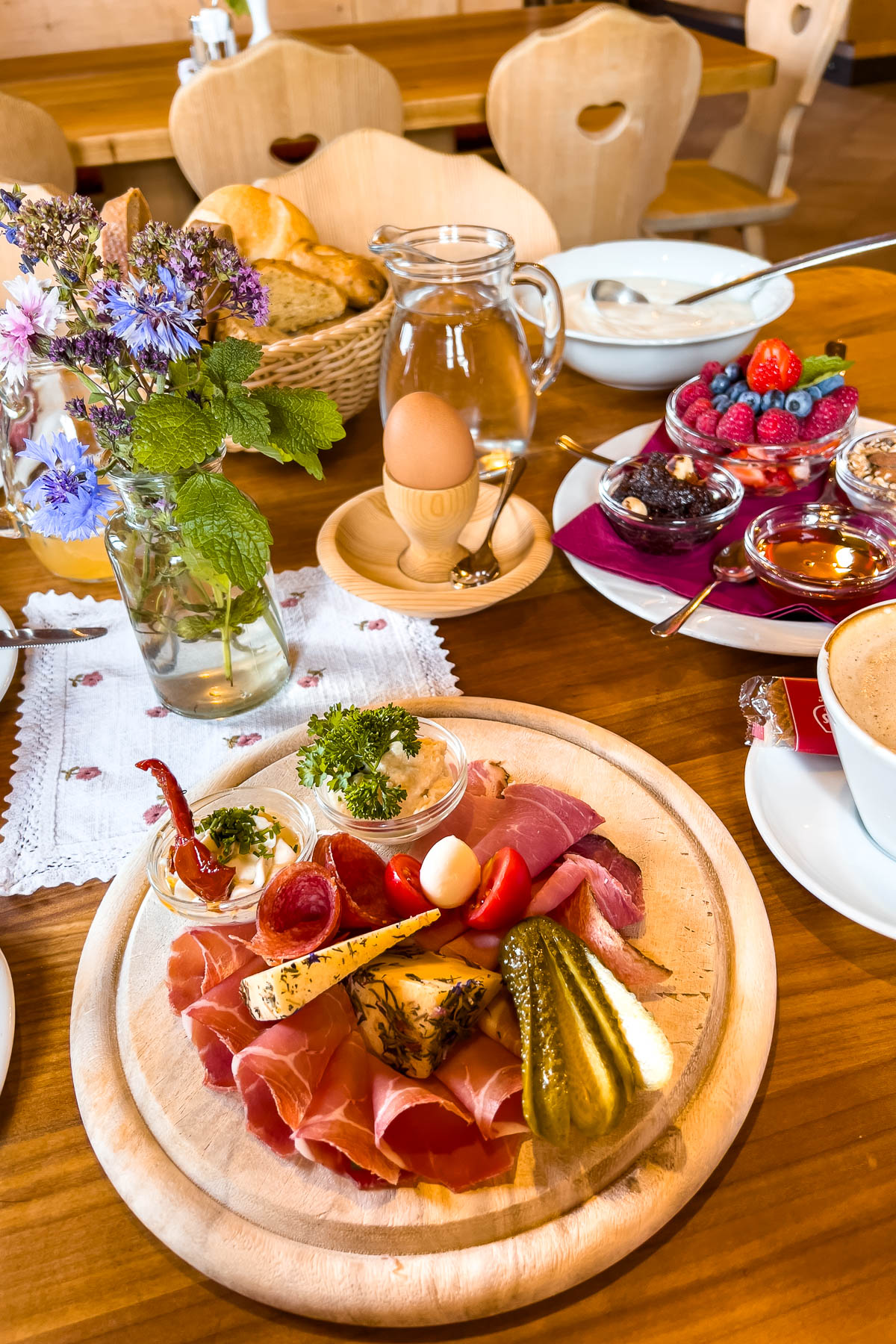A wooden table at Baita Troier Hütte at Seceda in the Dolomites with a traditional alpine breakfast spread including cured meats, cheese, bread, eggs, jam, berries, and coffee.