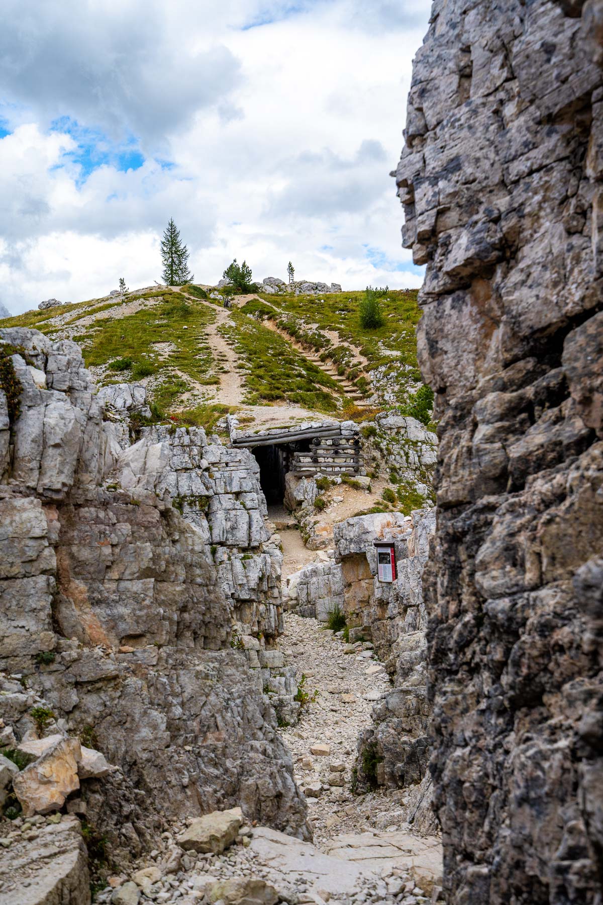 Rocky passage at Cinque Torri in the Dolomites, showing a narrow trail between stone walls leading toward old wartime bunkers and a hillside path above.