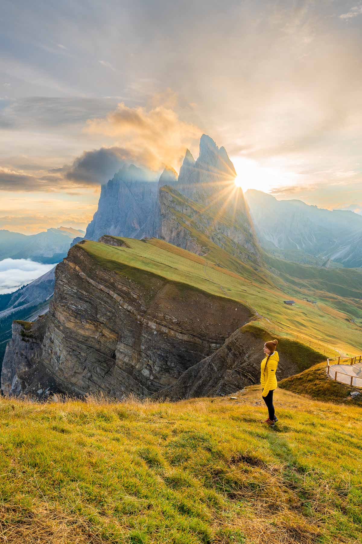 A stunning sunrise over Seceda, with golden light illuminating the grassy hills and the sharp peaks. A woman in a yellow jacket stands in the foreground, soaking in the view.