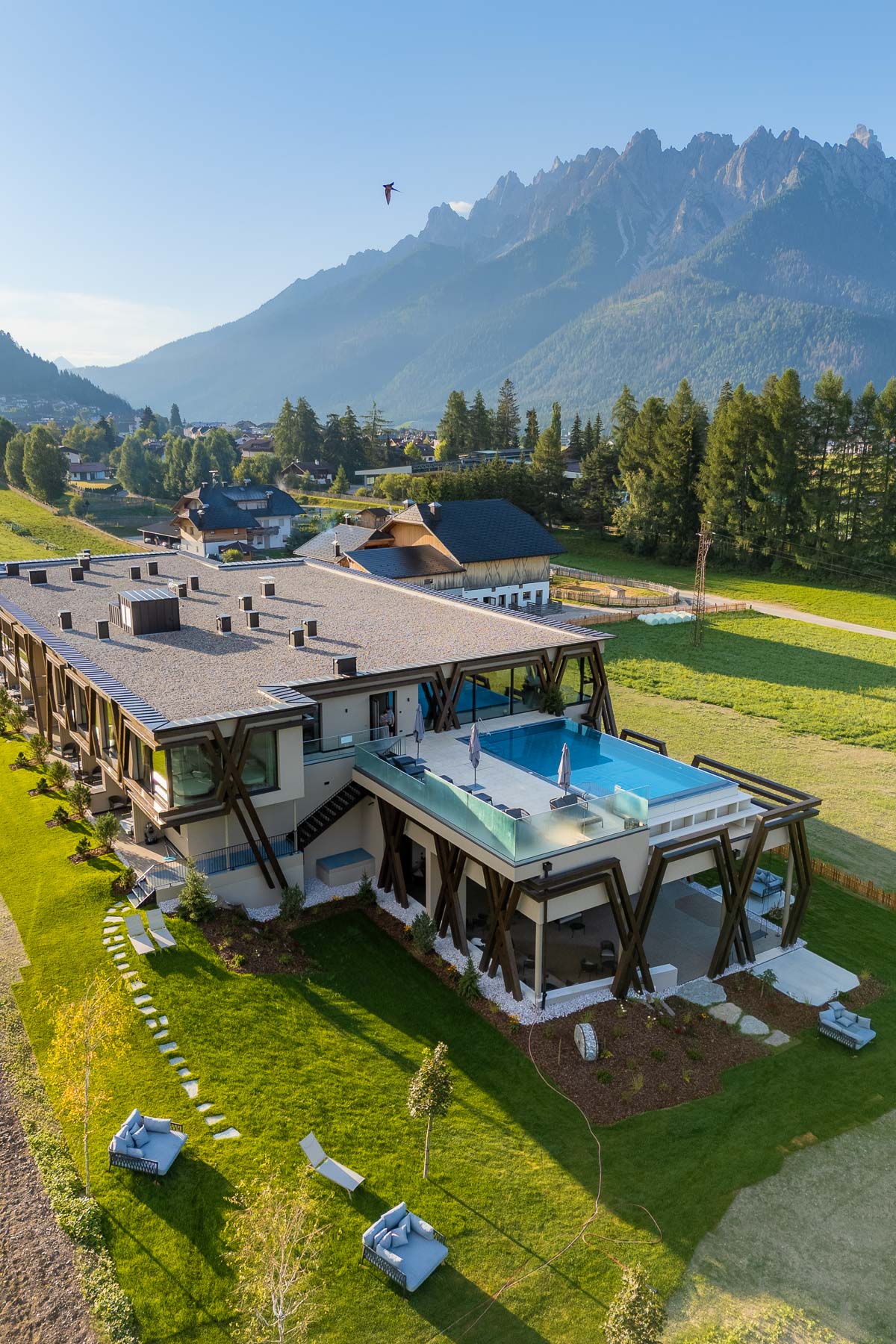 Aerial view of Hotel Patris in the Dolomites with an outdoor pool, modern alpine-style building, green lawns around it, and jagged mountain peaks rising in the background.