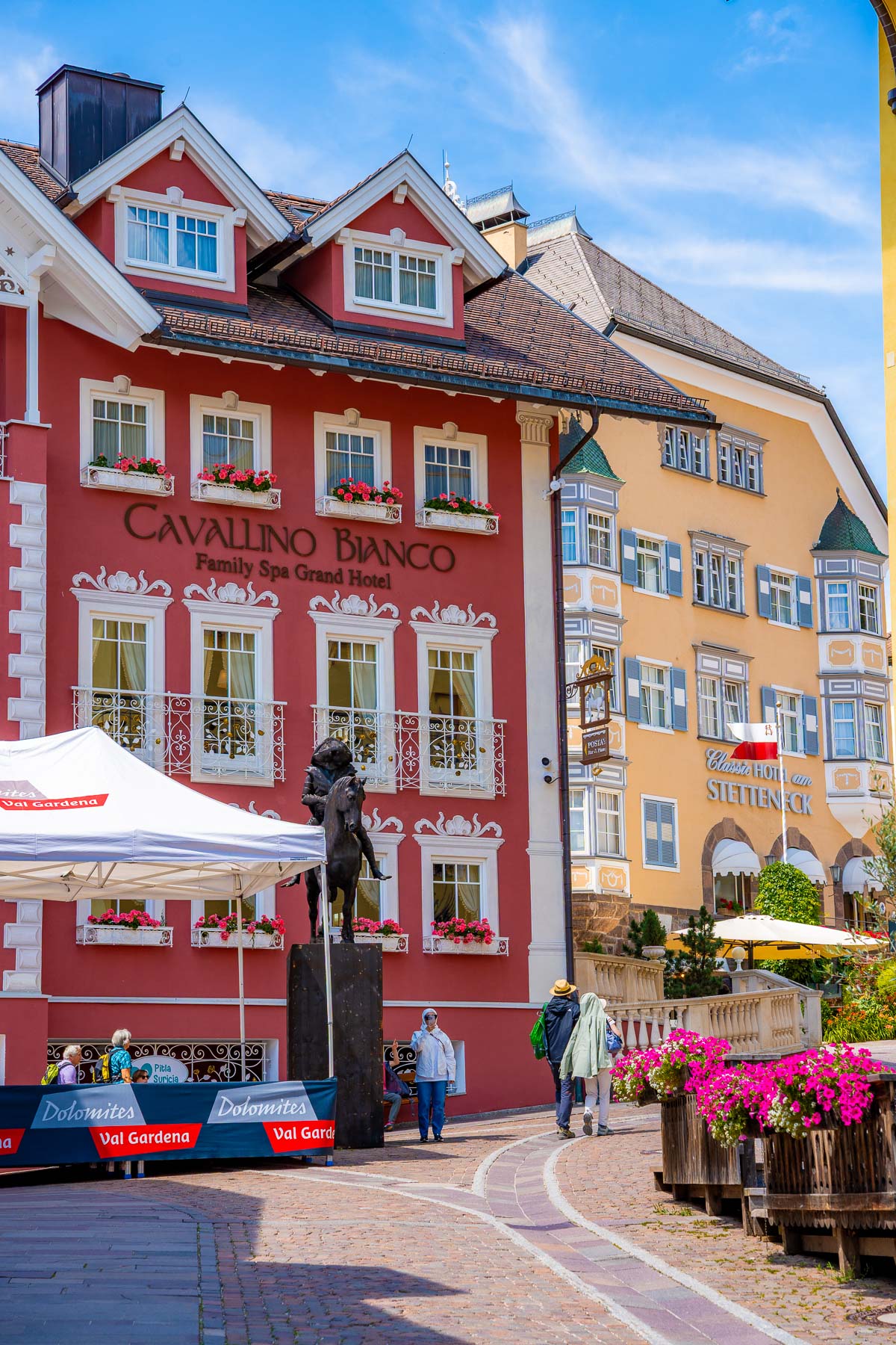 Colorful buildings lining a pedestrian street in Ortisei, Dolomites, with flower boxes on the windows, people walking through the square, and a statue set up in front of the red Cavallino Bianco building.