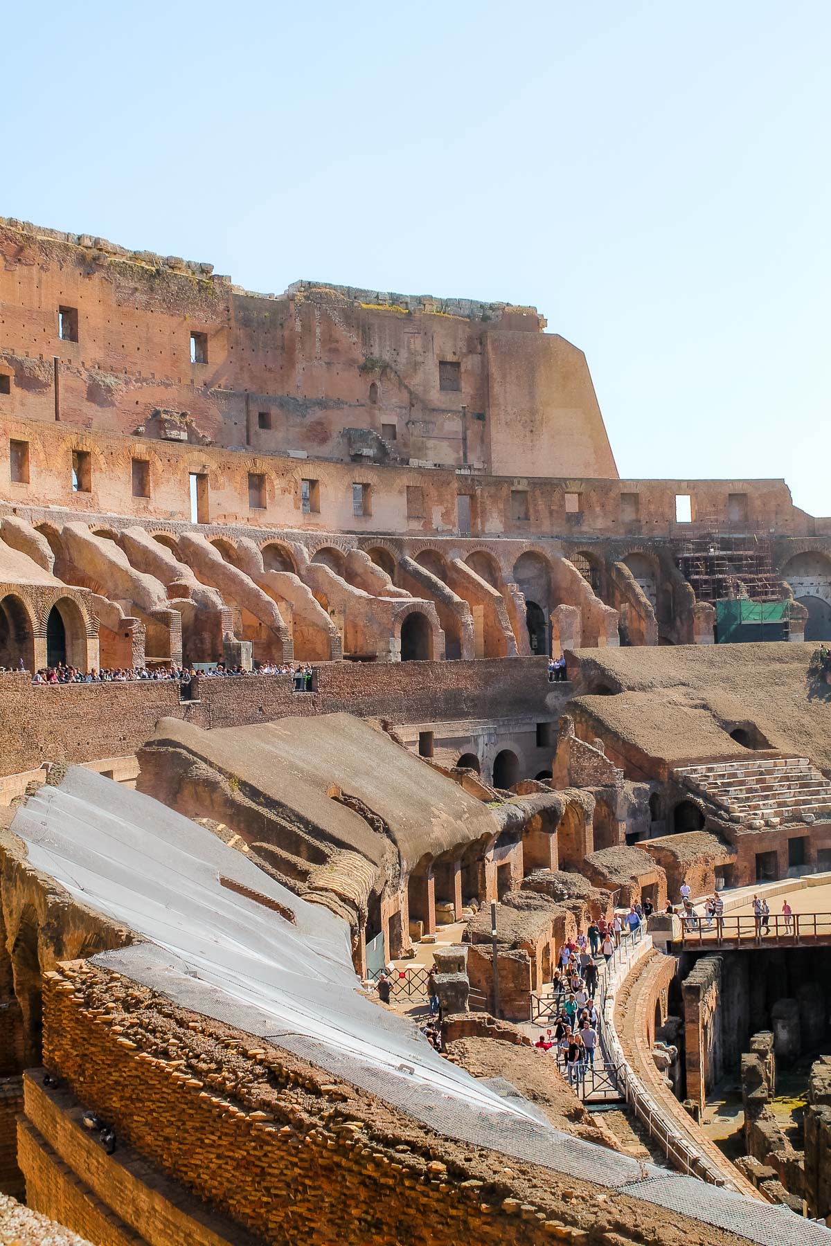 Interior view of the Colosseum in Rome, Italy, showing layered stone arches, walkways, and groups of visitors moving through the lower levels.