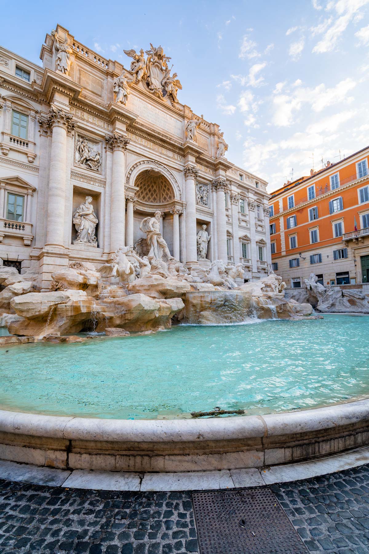 Wide view of the Trevi Fountain in Rome with turquoise water in the foreground, marble statues and columns behind it, and surrounding buildings framing the fountain.