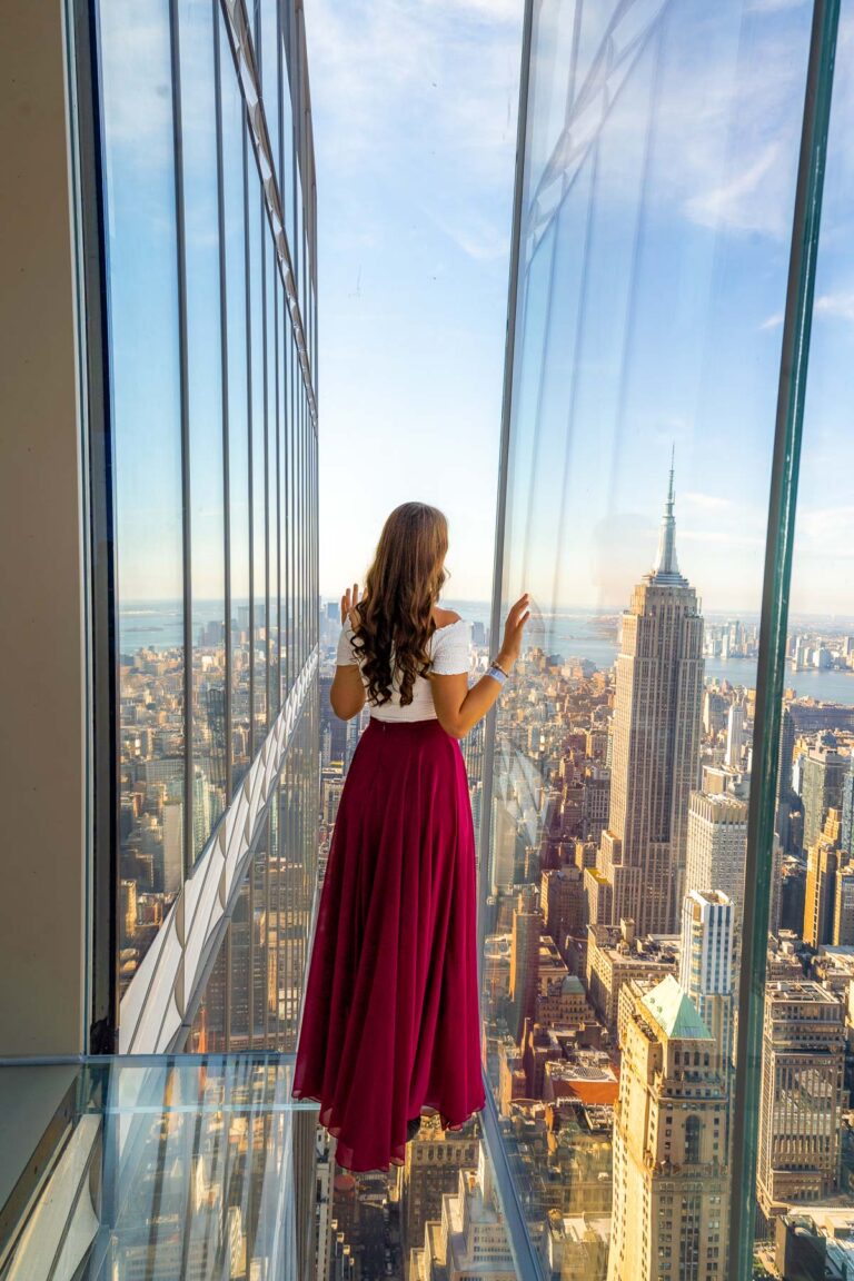 A person in a red skirt looking out from SUMMIT One Vanderbilt, with hands touching the glass walls. The Empire State Building and city skyline stretch out below.