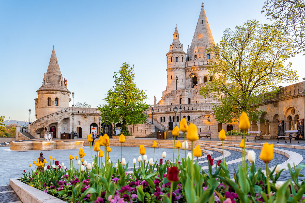 Fisherman’s Bastion in Budapest is shown with its towers and arches, with colorful flowers in the foreground.