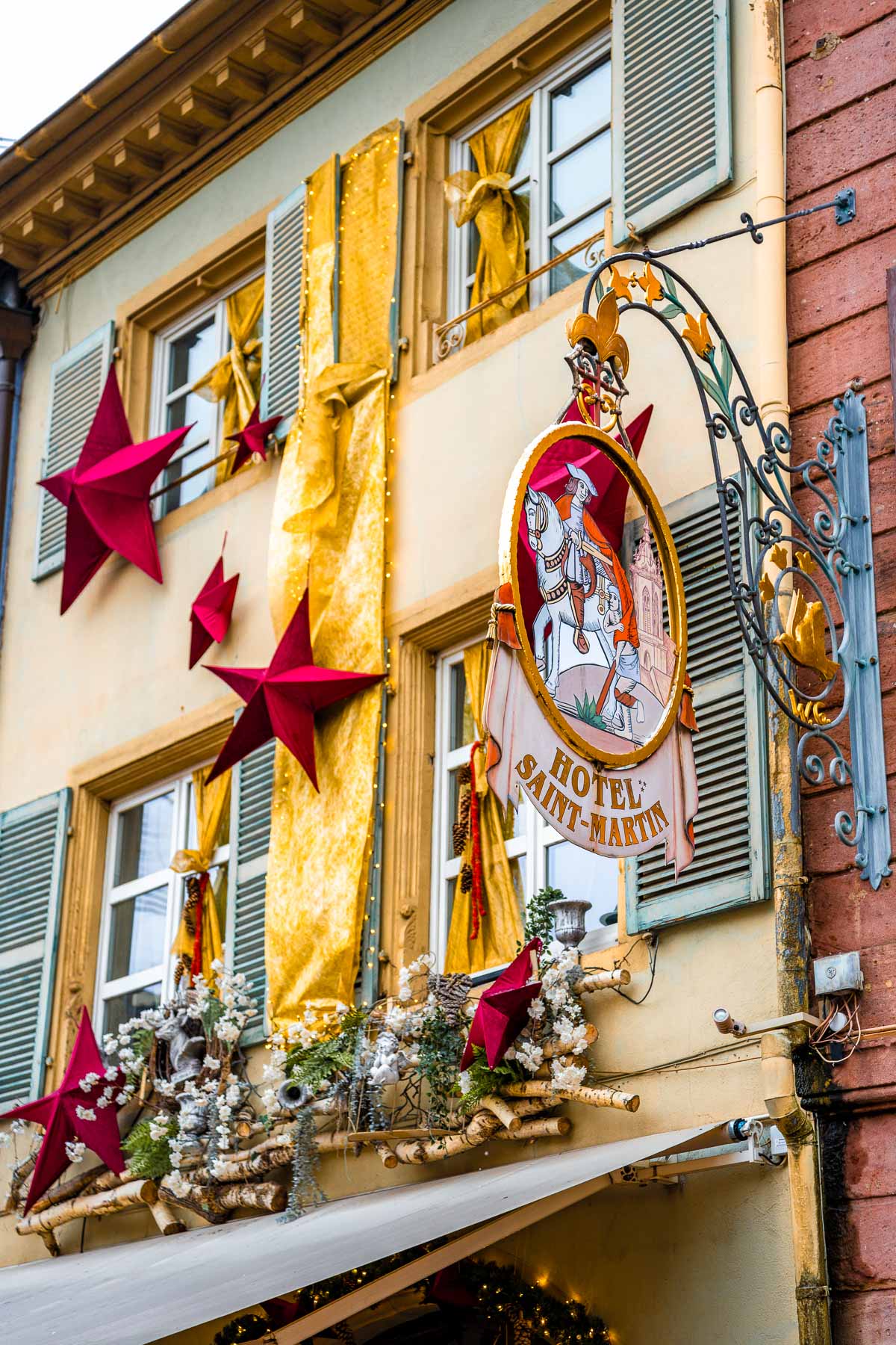 The sign for Hôtel Saint-Martin in Colmar, France hangs on a decorative metal bracket beside shuttered windows and gold fabric bows. Red star decorations and garlands are displayed across the building’s front.
