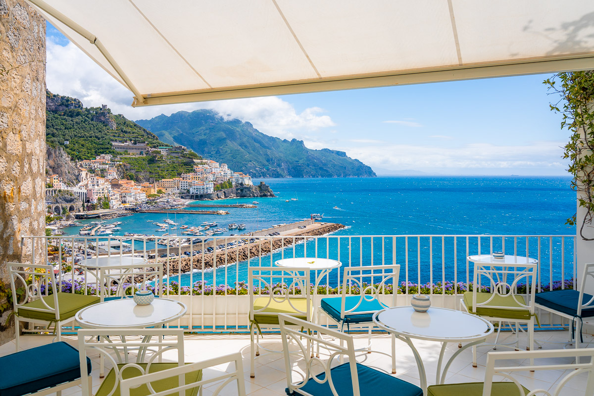 The terrace of Mario's Lounge Bar at Hotel Miramalfi in Amalfi, with white tables and chairs overlooking the coastline, harbor, and turquoise waters of the Amalfi Coast.