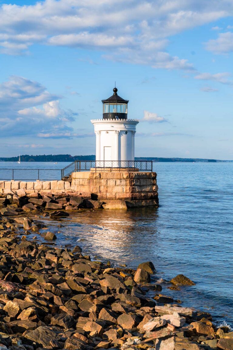 A small white lighthouse, Bug Light in Portland, Maine, with a black top stands at the end of a rocky breakwater, surrounded by calm blue water. In the background, a shoreline with buildings and sailboats is visible under a partly cloudy sky.