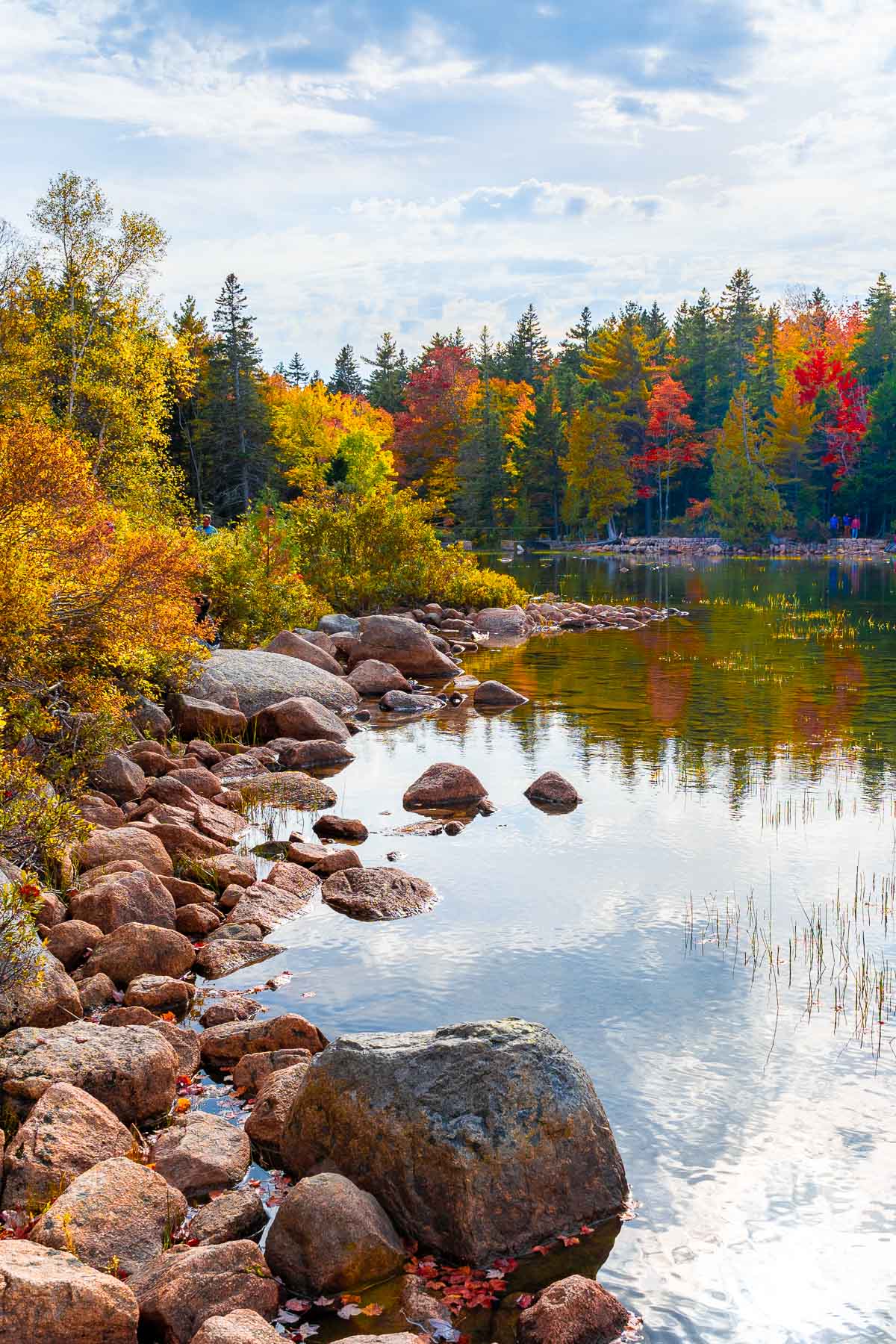 The peaceful Jordan Pond in Acadia National Park with crystal-clear water reflecting the autumn colors of the surrounding trees. Large rocks line the edge of the pond.