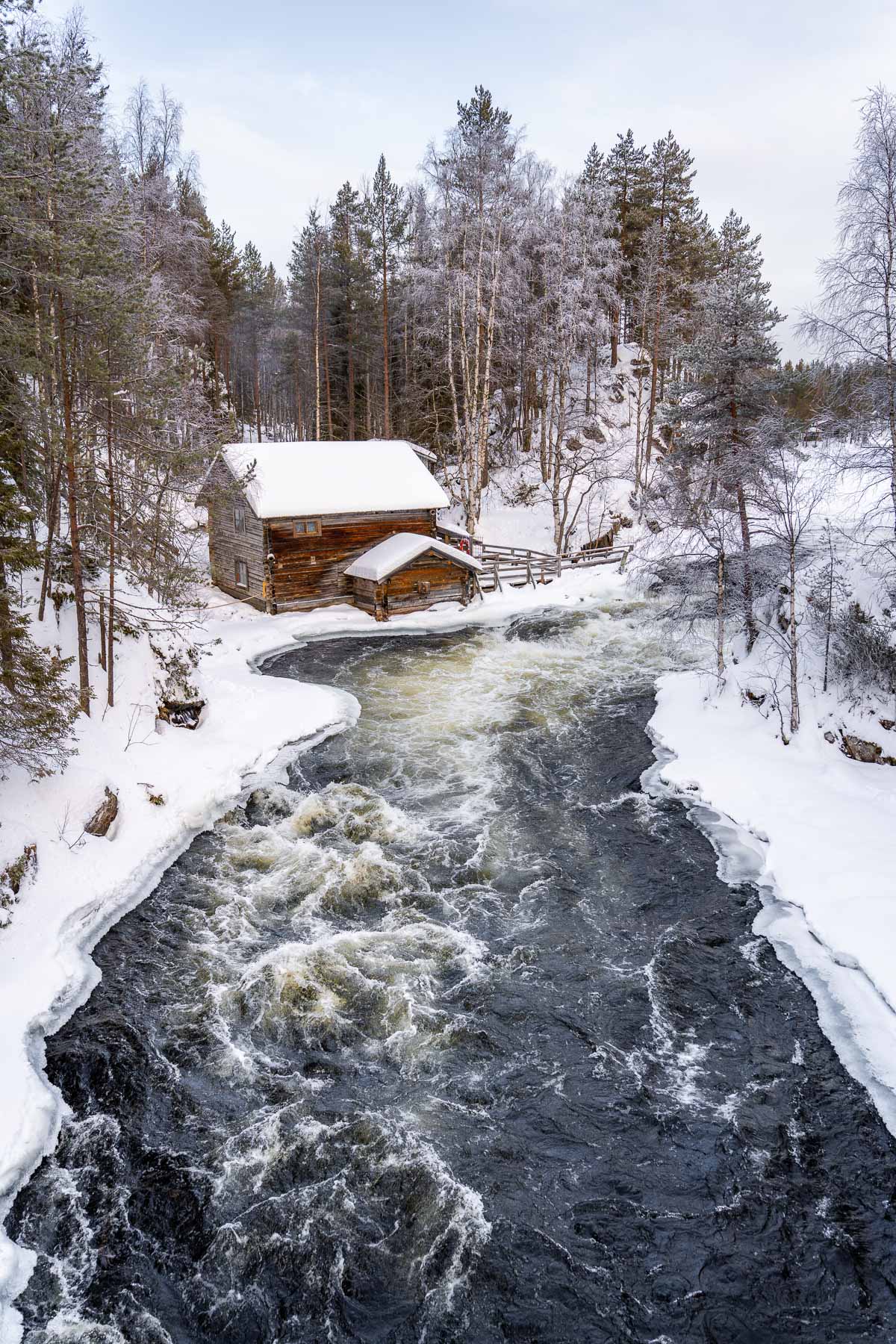 A wooden cabin with a snow-covered roof sits beside the rushing Myllykoski Rapids in Oulanka National Park, Lapland, surrounded by frosty trees and winter scenery.
