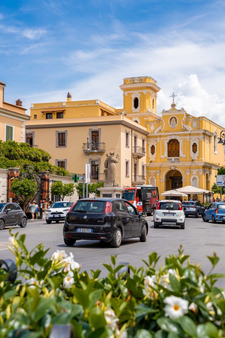 Piazza Tasso in Sorrento, featuring vibrant yellow buildings, a central statue, and cars and people passing through the bustling square on a sunny day.