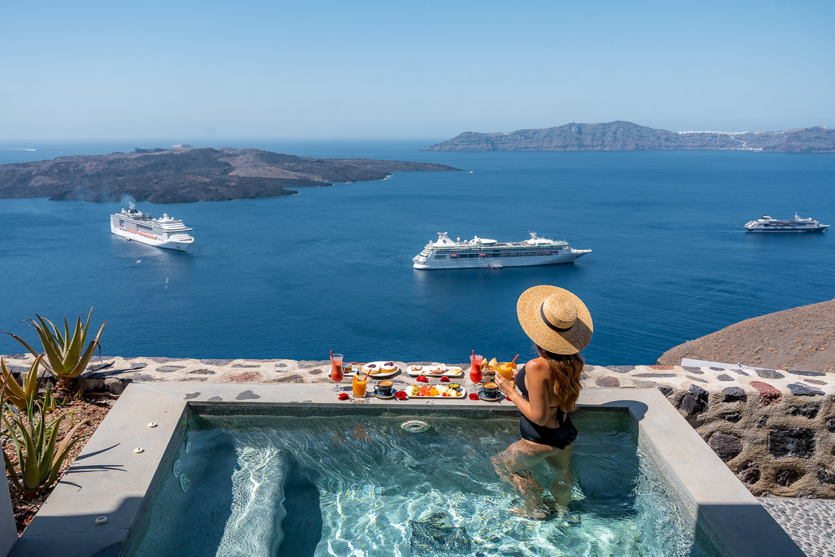 A woman in a black swimsuit and wide-brimmed straw hat enjoying a floating breakfast in a private pool at Villa Bordeaux in Santorini. The stunning view showcases the caldera and cruise ships on the Aegean Sea under a clear blue sky.