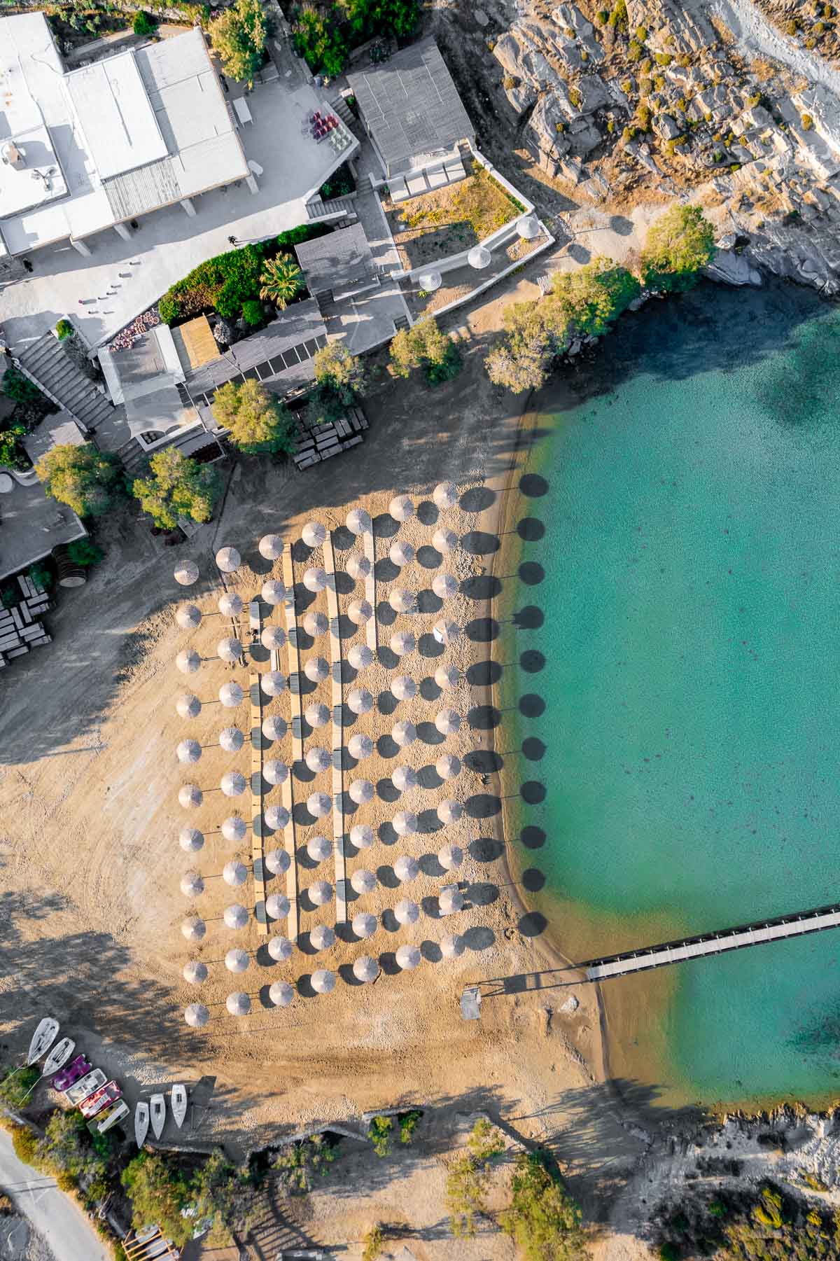 Aerial view of Monastiri Beach, one of the best beaches in Paros, featuring rows of umbrellas on golden sand, turquoise waters, and nearby facilities nestled among lush greenery and rocky cliffs.