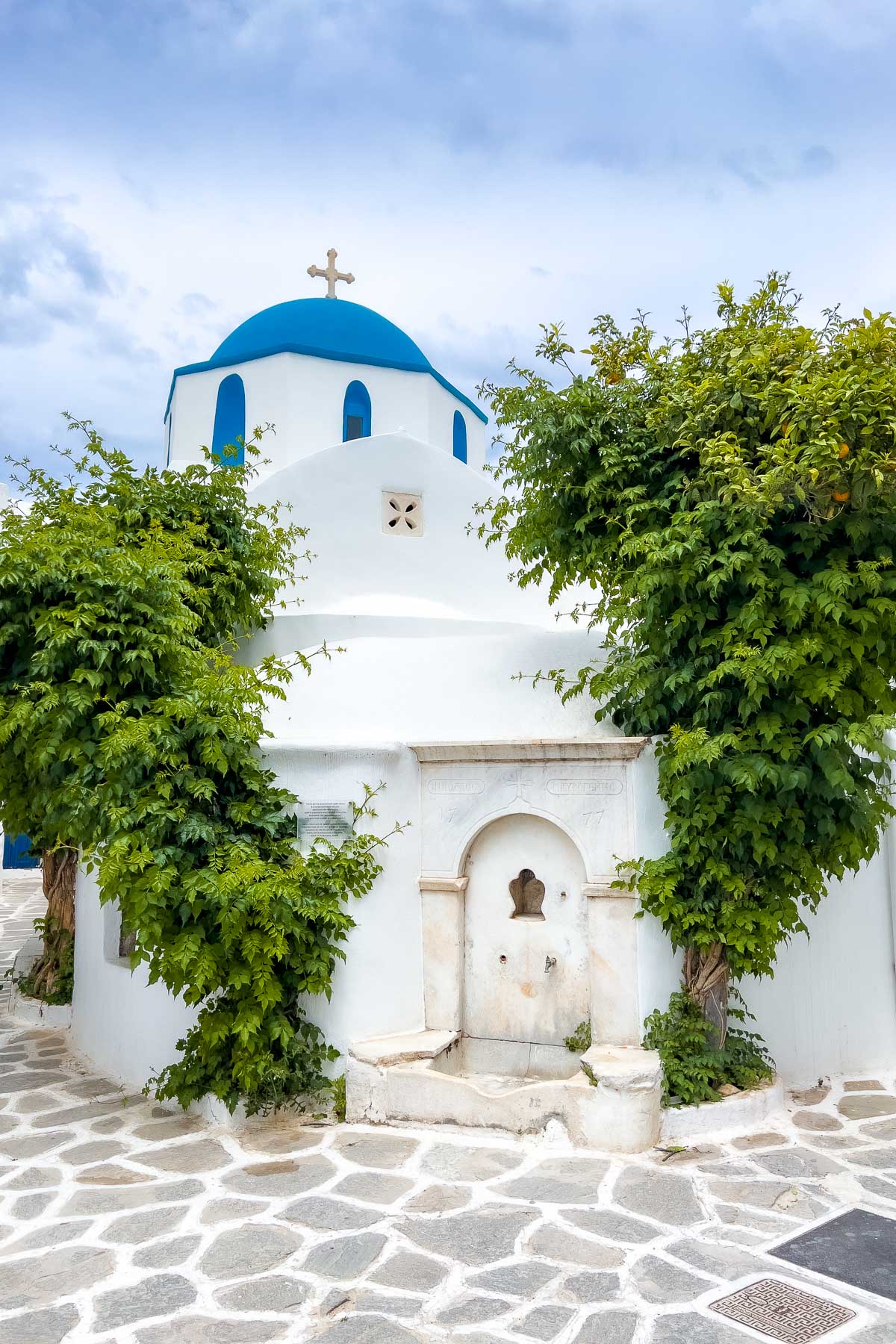 The iconic blue-domed church of Parikia is surrounded by lush green ivy. Its crisp white walls and cobblestone courtyard reflect the traditional beauty of Cycladic architecture.