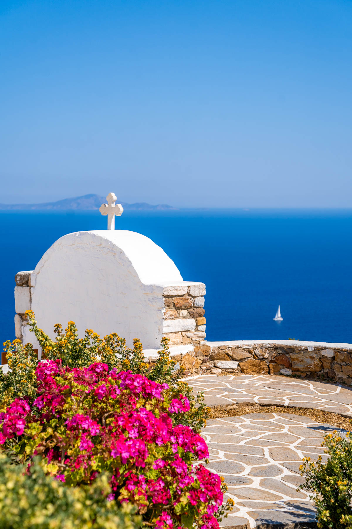 The rooftop cross of St. Antonios Monastery in Paros rises against a bright blue sky, with a sailboat floating in the distant Aegean Sea and pink flowers in the foreground.