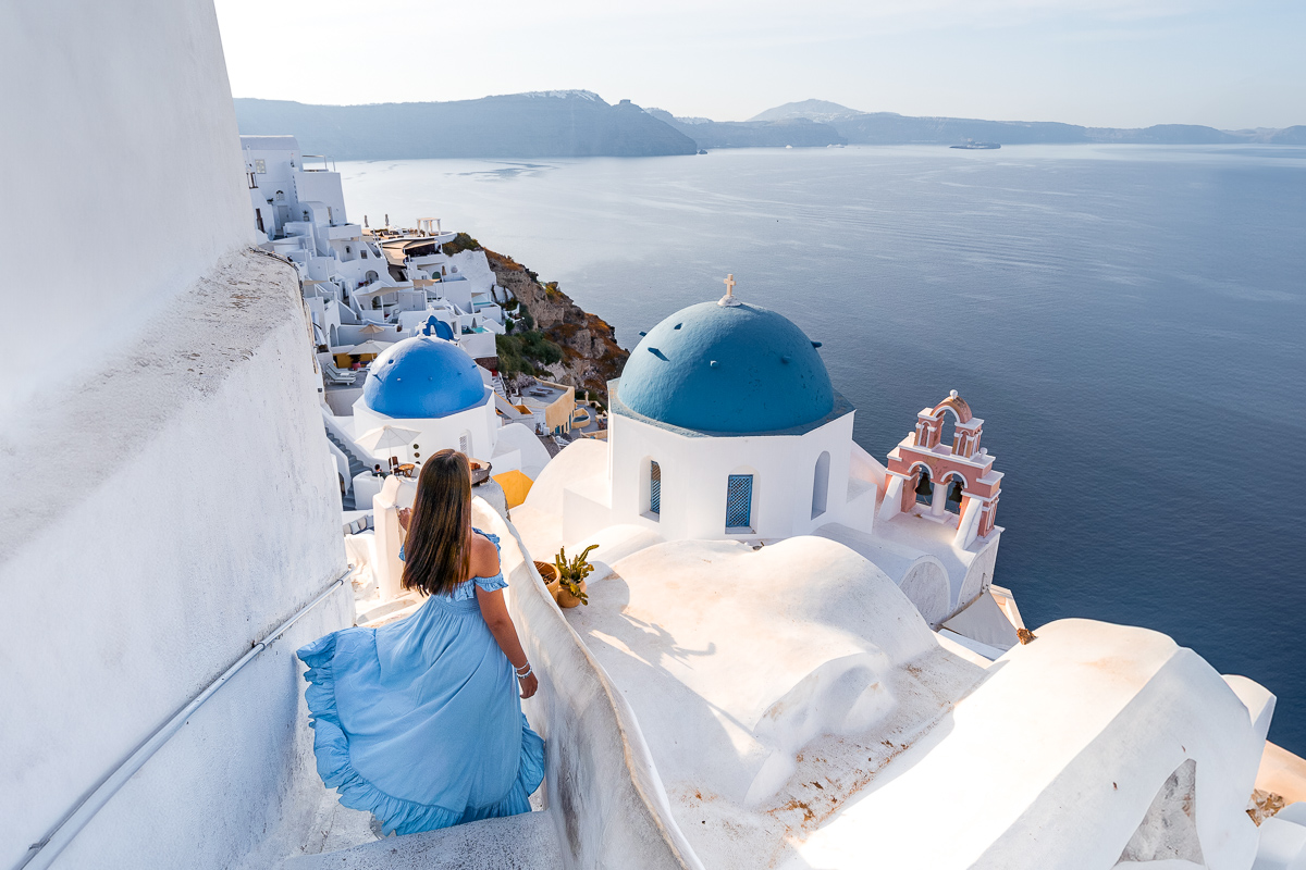 A woman in a light blue dress walks down a set of white stairs in Oia, Santorini. The three blue domes of the churches that are known as one of the best Santorini Instagram spots are visible to her right, and the caldera stretches out into the distance with calm waters.