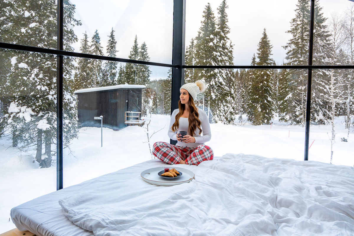 A cozy morning inside a glass igloo at Magical Pond, Lapland. A woman sits on a bed with white linens, wearing a beanie and plaid pajama pants, holding a cup of coffee. A plate of pastries rests beside her as she looks out at the snowy landscape through the floor-to-ceiling windows. Snow-covered trees and another igloo cabin are visible outside.