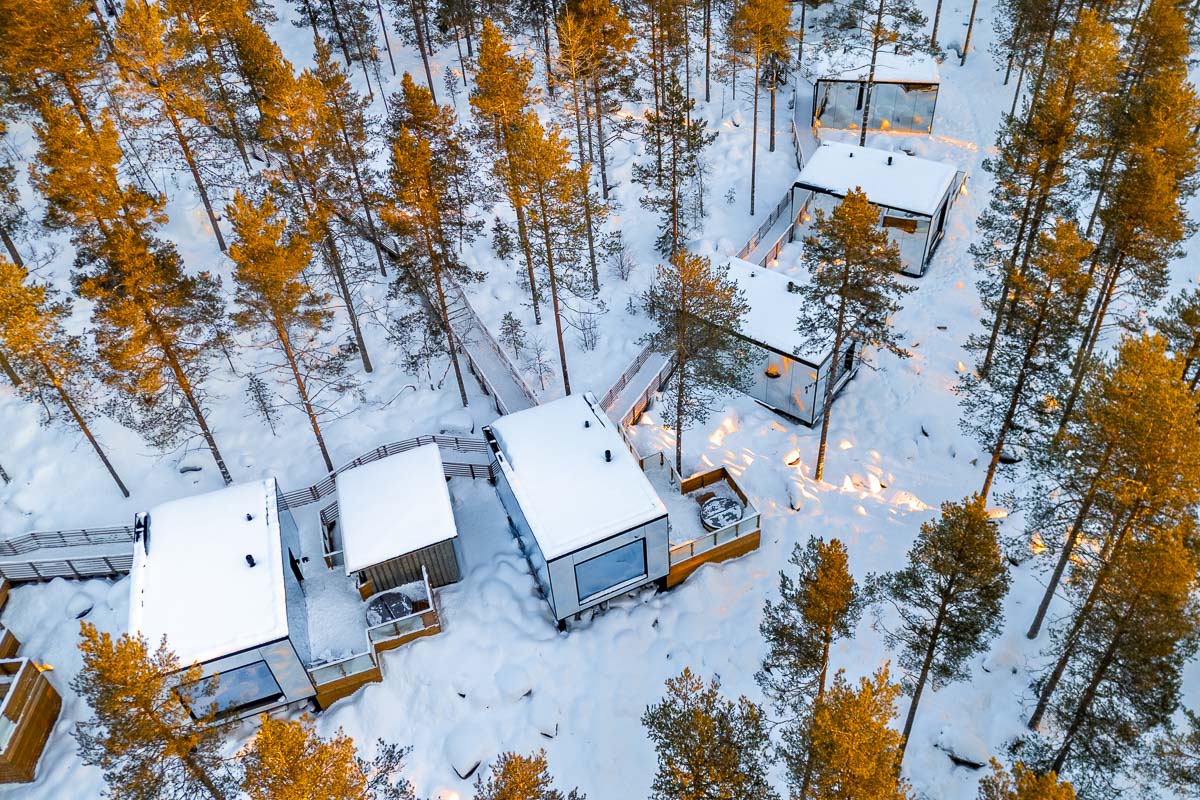 Aerial view of the Invisible Forest Lodge in Lapland, showing modern glass-walled cabins surrounded by snow-covered pine trees during golden hour.