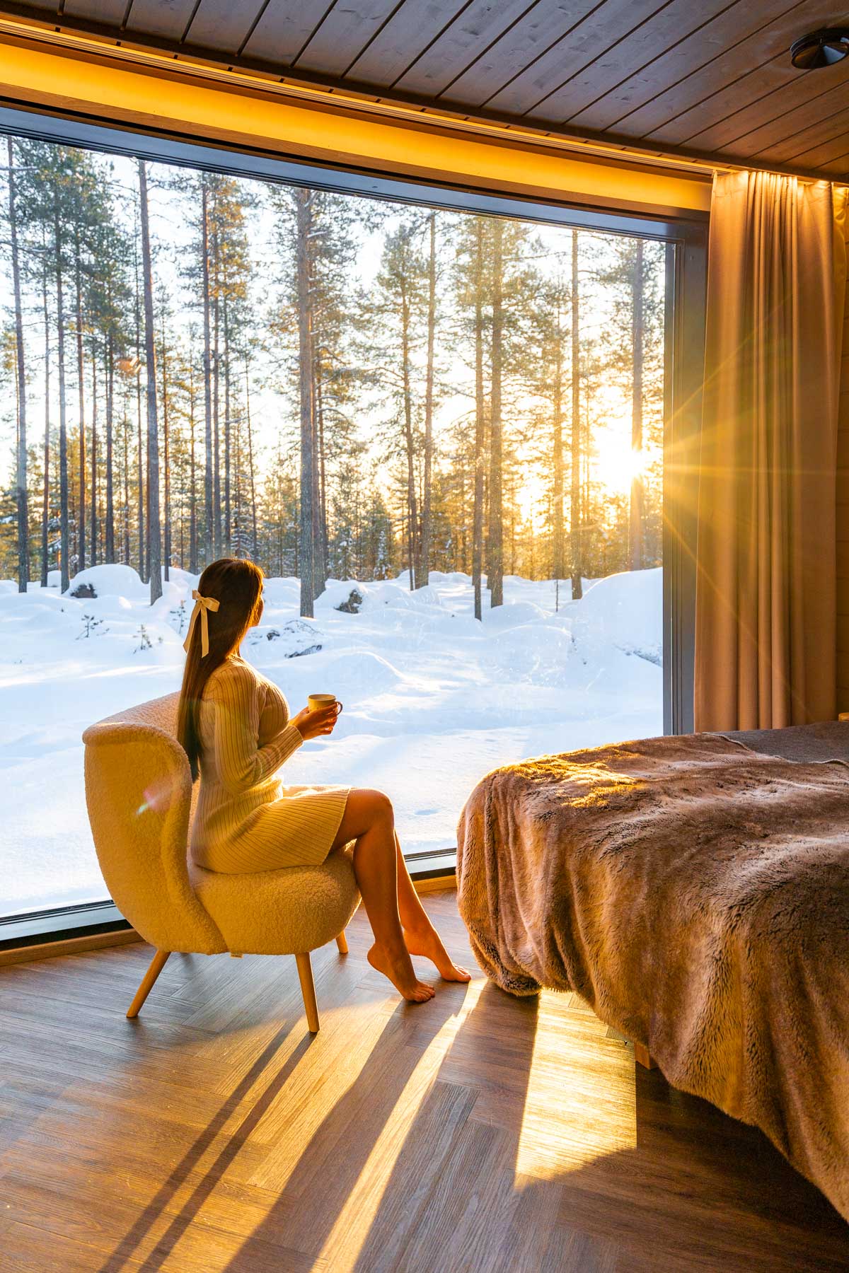 A woman sits in a cozy chair inside the Invisible Suite at Invisible Forest Lodge, holding a cup and looking out at the snow-covered forest through a floor-to-ceiling window as the sun rises.