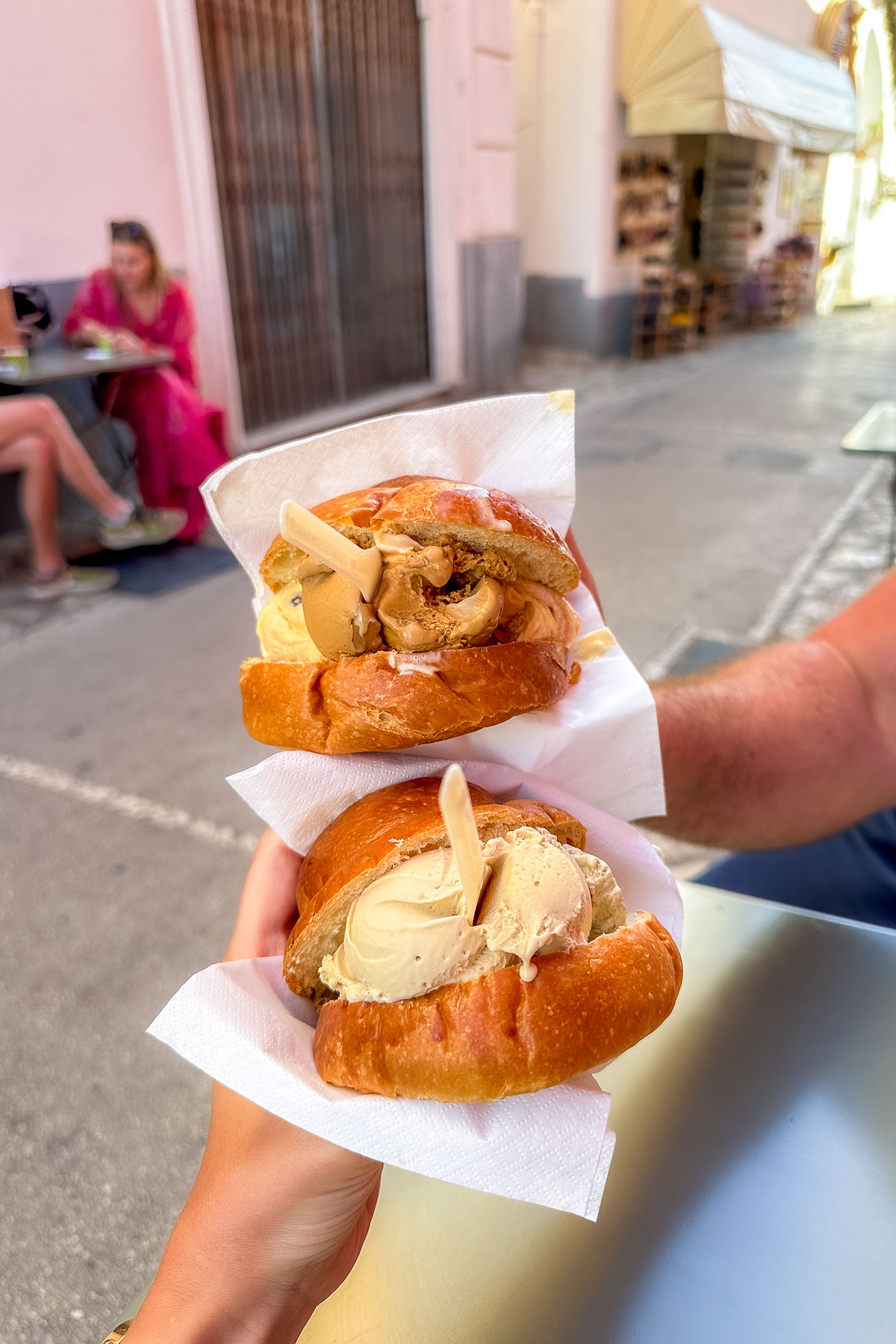 Two brioche buns filled with gelato, held up in an outdoor seating area at Capri Crema Cafe. The background shows a blurred street scene with people sitting at tables and a shop with an awning.