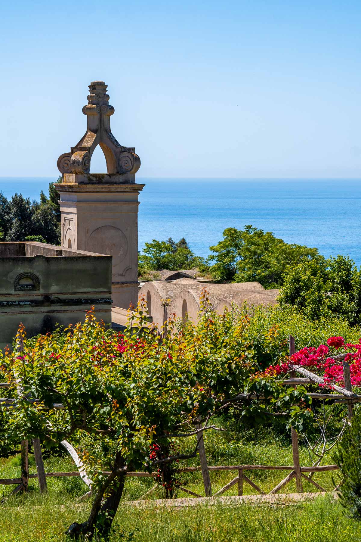 The historic stone structure with an ornate arched detail of Certosa San Giacomo in Capri, surrounded by lush greenery and red flowers, with the blue sea visible in the background.
