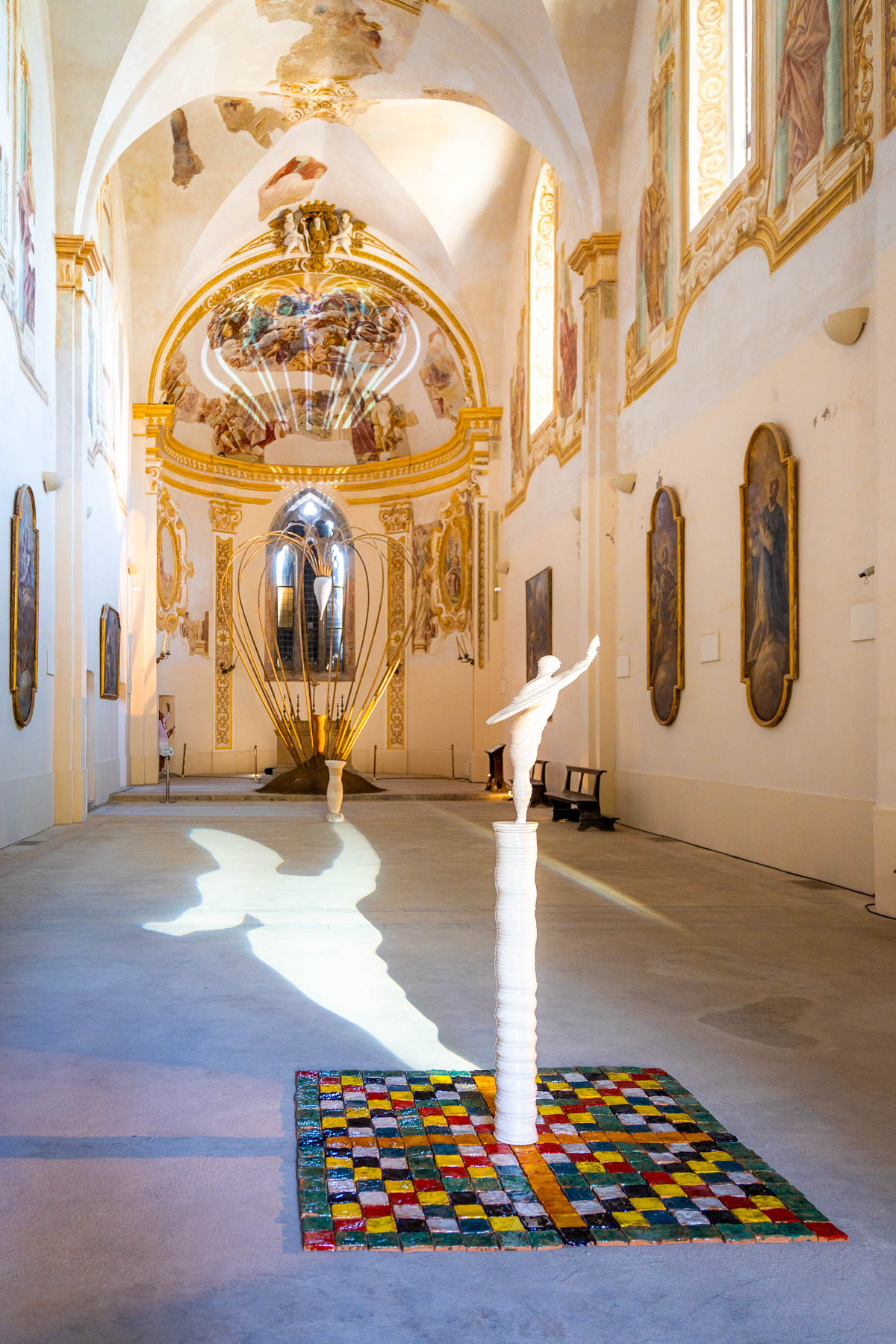 The interior of Certosa San Giacomo in Capri, featuring high vaulted ceilings with frescoes, golden details, and a modern sculpture in the center casting a shadow on the floor.