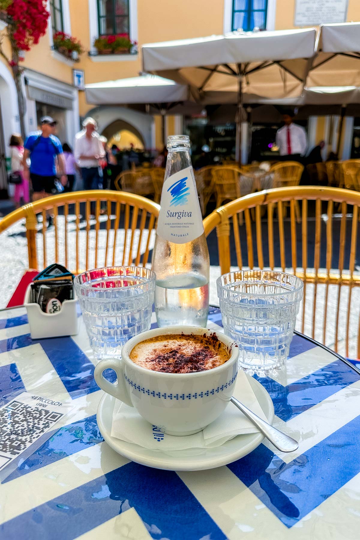 A close-up of a cappuccino served in a white cup with blue detailing, placed on a checkered blue-and-white table at Il Piccolo in Capri. A glass bottle of Surgiva water and two glasses are also on the table, with outdoor seating and people walking in the background.