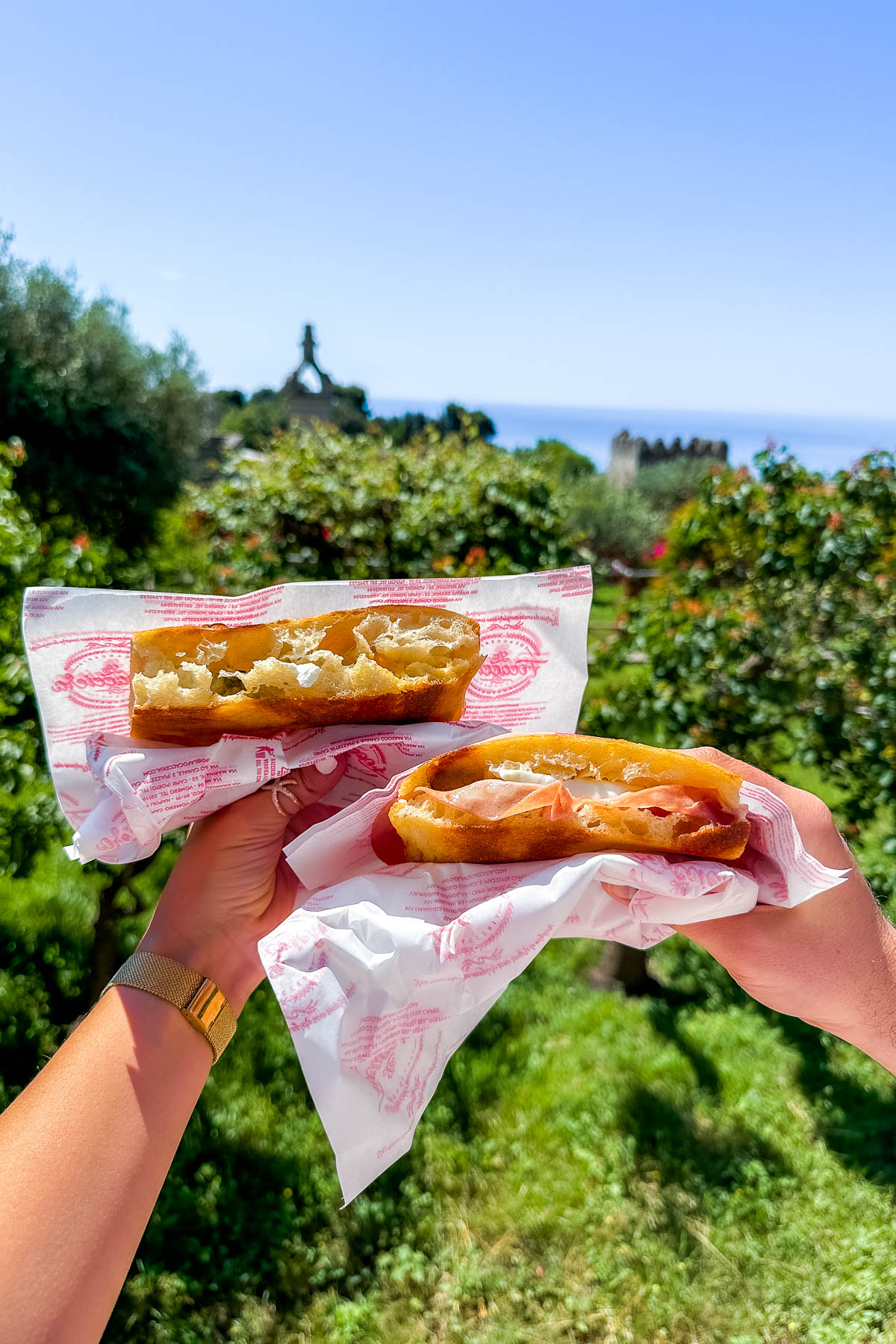 Two hands holding fresh focaccia sandwiches from La Focacciera in Capri. The background shows greenery, a stone statue, and a glimpse of the sea.