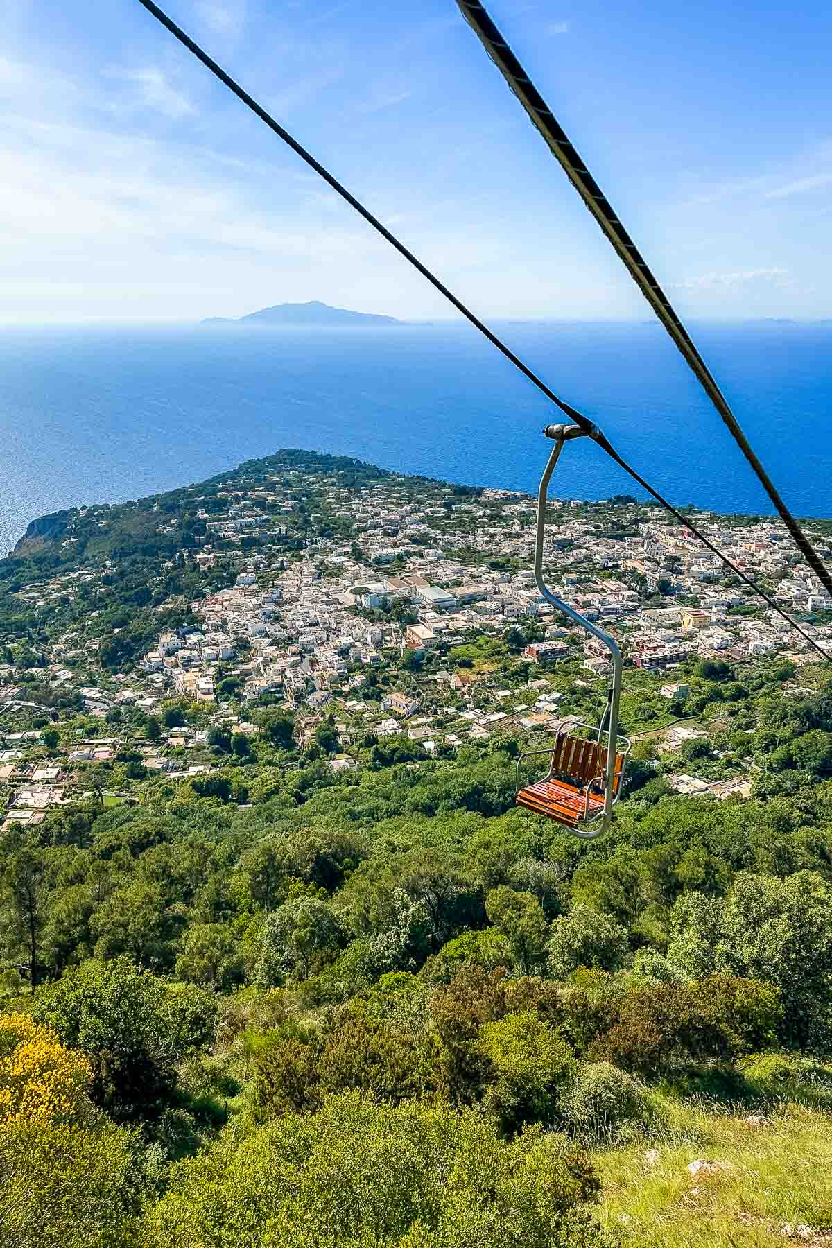 A single-seat chairlift suspended above lush green hills, offering a view of Anacapri, the blue sea, and distant islands in the background from Monte Solaro.