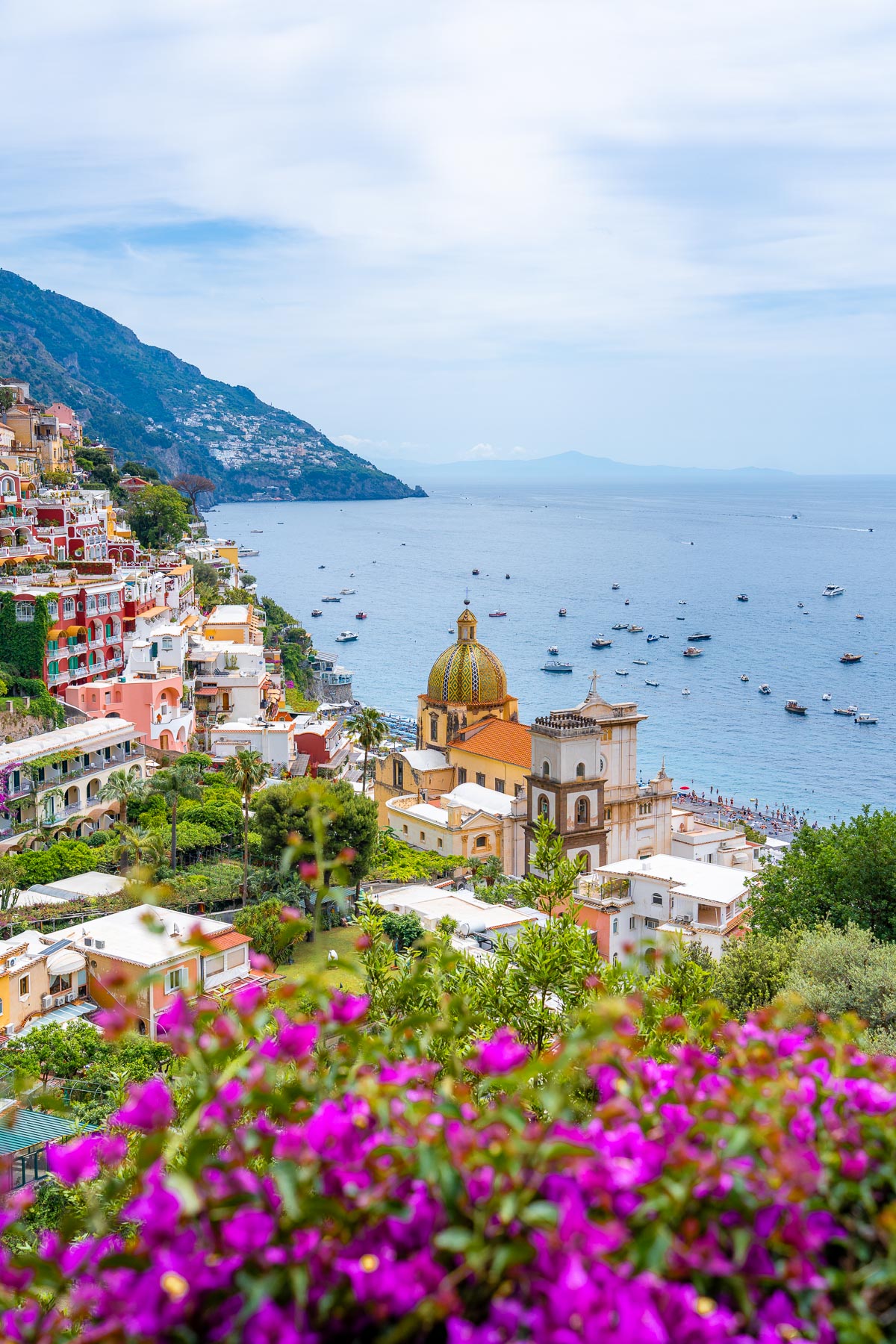 A scenic view of Positano, Italy, with the Church of Santa Maria Assunta's colorful dome in the foreground, vibrant pink flowers, and boats dotting the blue waters of the Amalfi Coast.