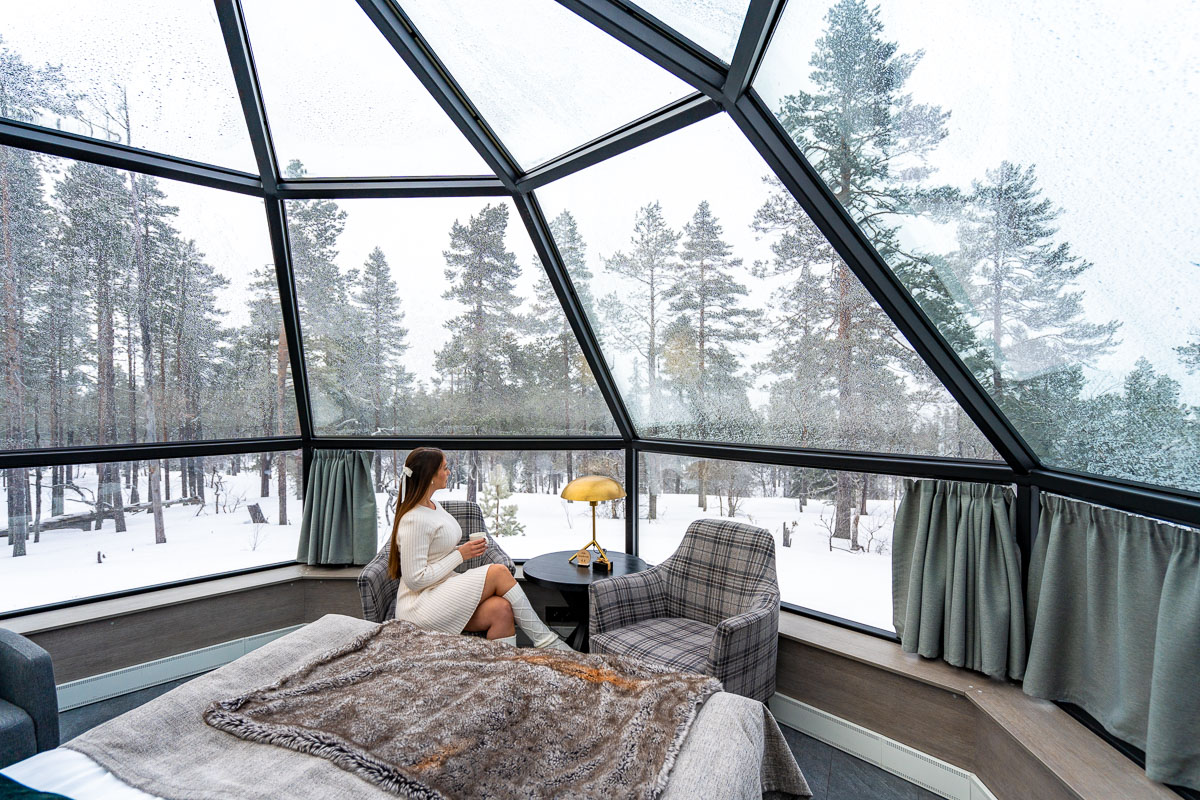 Inside a Prime Superior Igloo at Levin Iglut, a woman in a white sweater dress sits by the window with a drink, looking at the snow-covered forest outside. The glass walls provide a full view of the winter landscape, with frost forming on the windows.