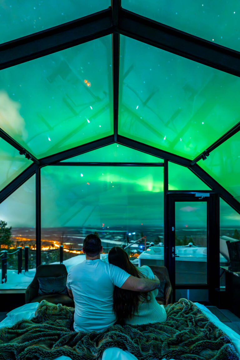 A couple sits on a bed inside a glass igloo at Levin Iglut, looking up at the Northern Lights. The green aurora is visible through the glass ceiling, with a snowy landscape and distant lights outside.