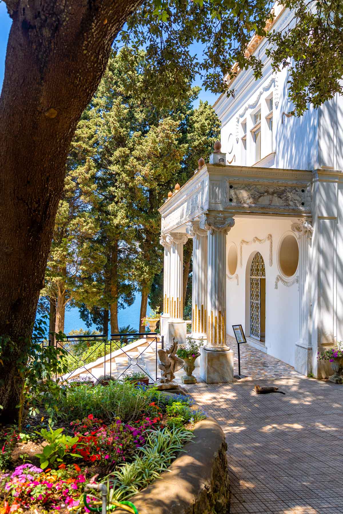 A white neoclassical villa with golden accents at Villa Lysis in Capri, surrounded by lush greenery and overlooking the sea. A stone pathway leads to the entrance, framed by colorful flowers.