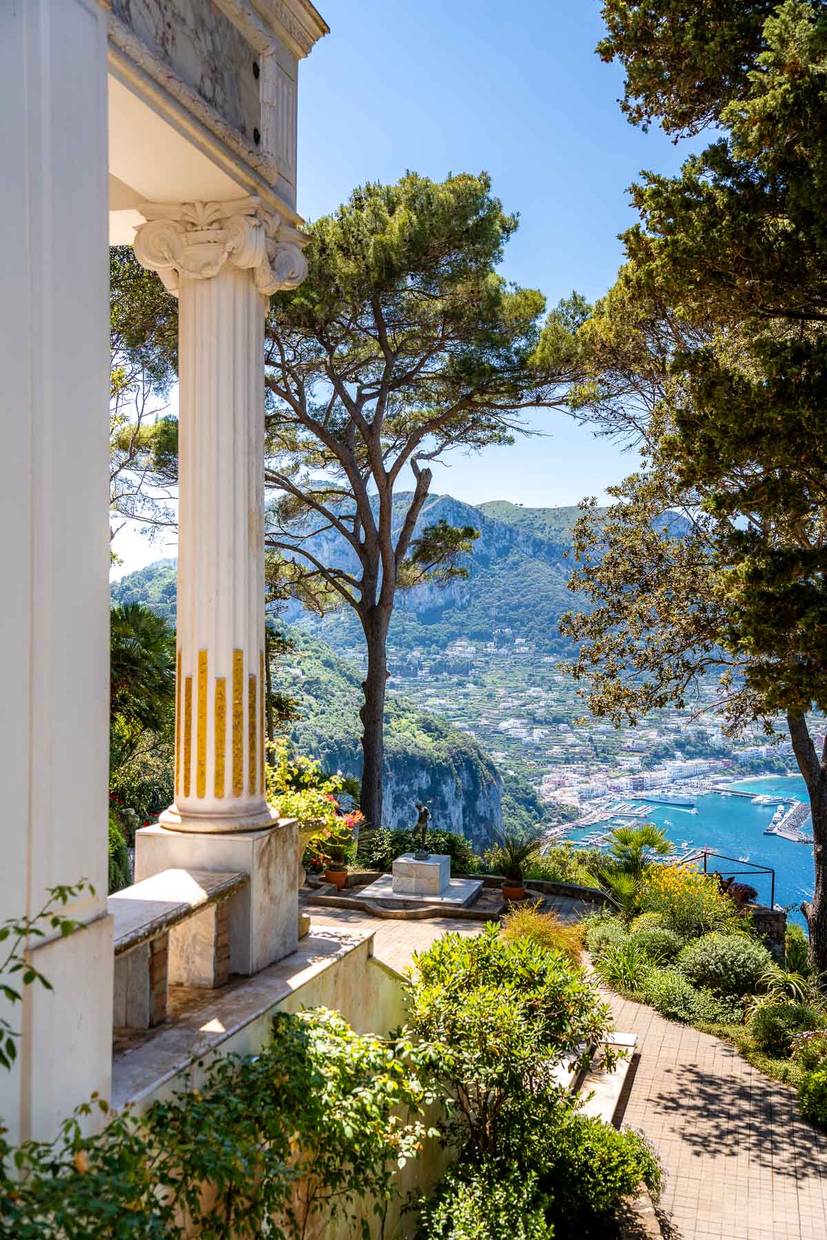 A scenic view from Villa Lysis in Capri, looking over the island’s cliffs, greenery, and Marina Grande in the distance. A classical column stands in the foreground, partially framing the landscape.