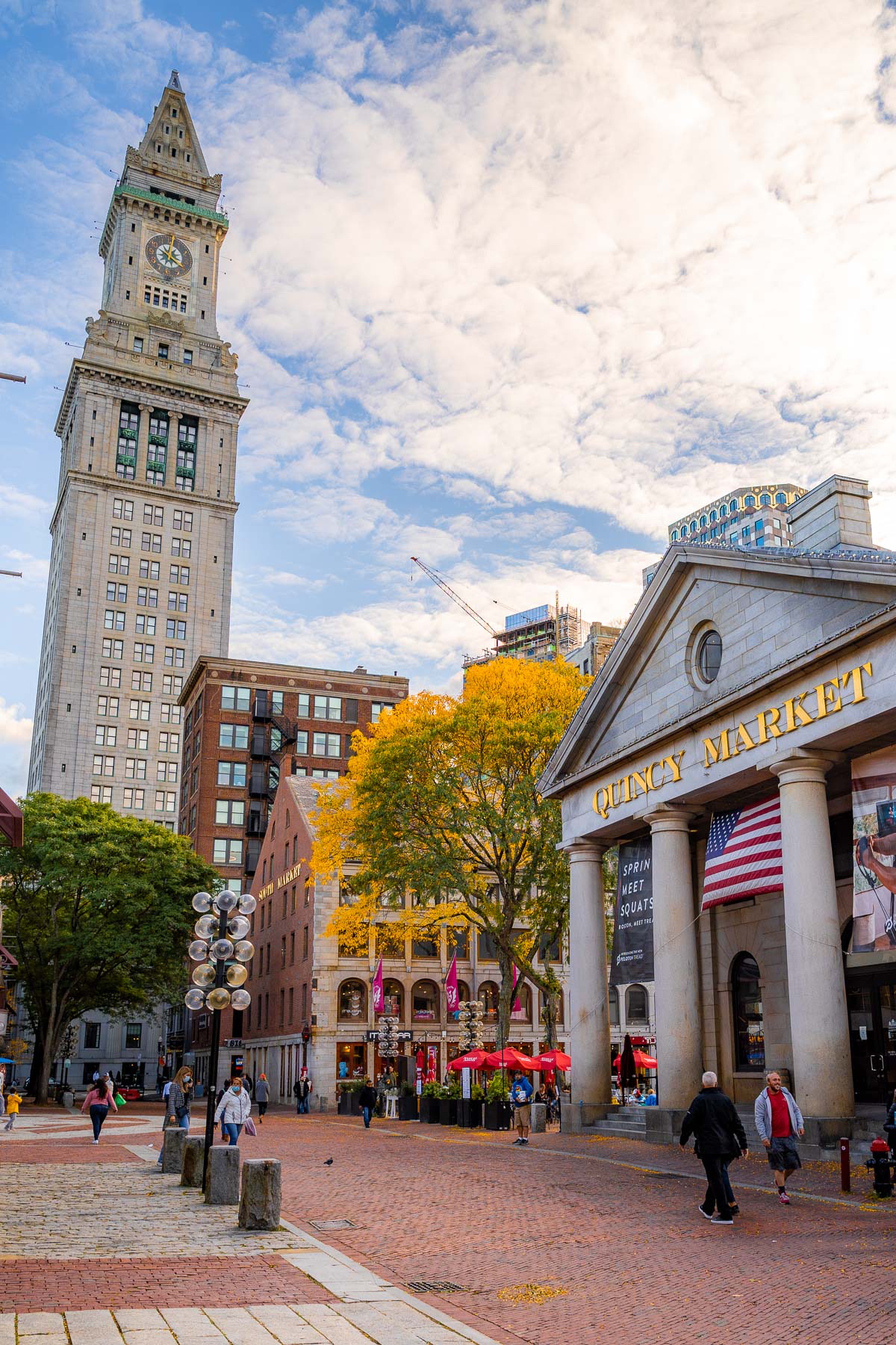 People walking around Quincy Market in Boston, with the historic building on the right, fall trees behind it, and the Custom House Tower visible in the background.