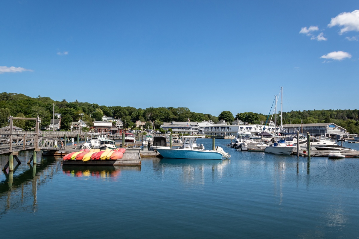 Docked boats and a bunch of bright kayaks floating near the piers in Boothbay Harbor, Maine, with waterfront buildings and greenery in the distance.
