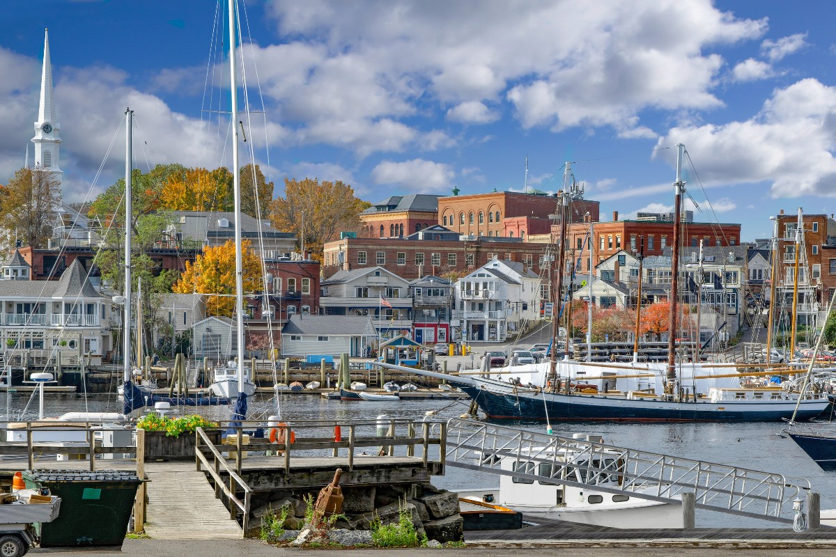 View of Camden, Maine, from the harbor with sailboats docked in the foreground and red brick buildings and a white steeple rising behind the waterfront homes and shops.