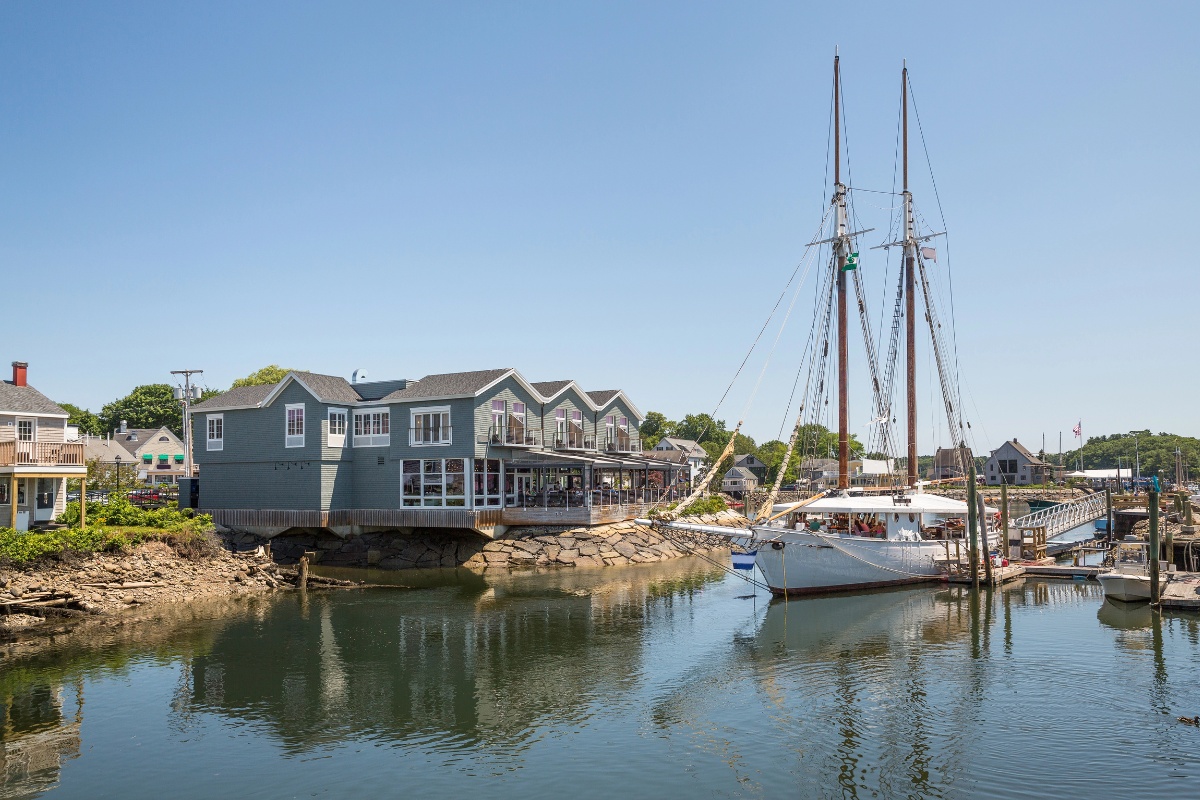 A sailboat docked in the harbor in Kennebunkport, Maine, next to a gray waterfront building with large windows and balconies overlooking the water.
