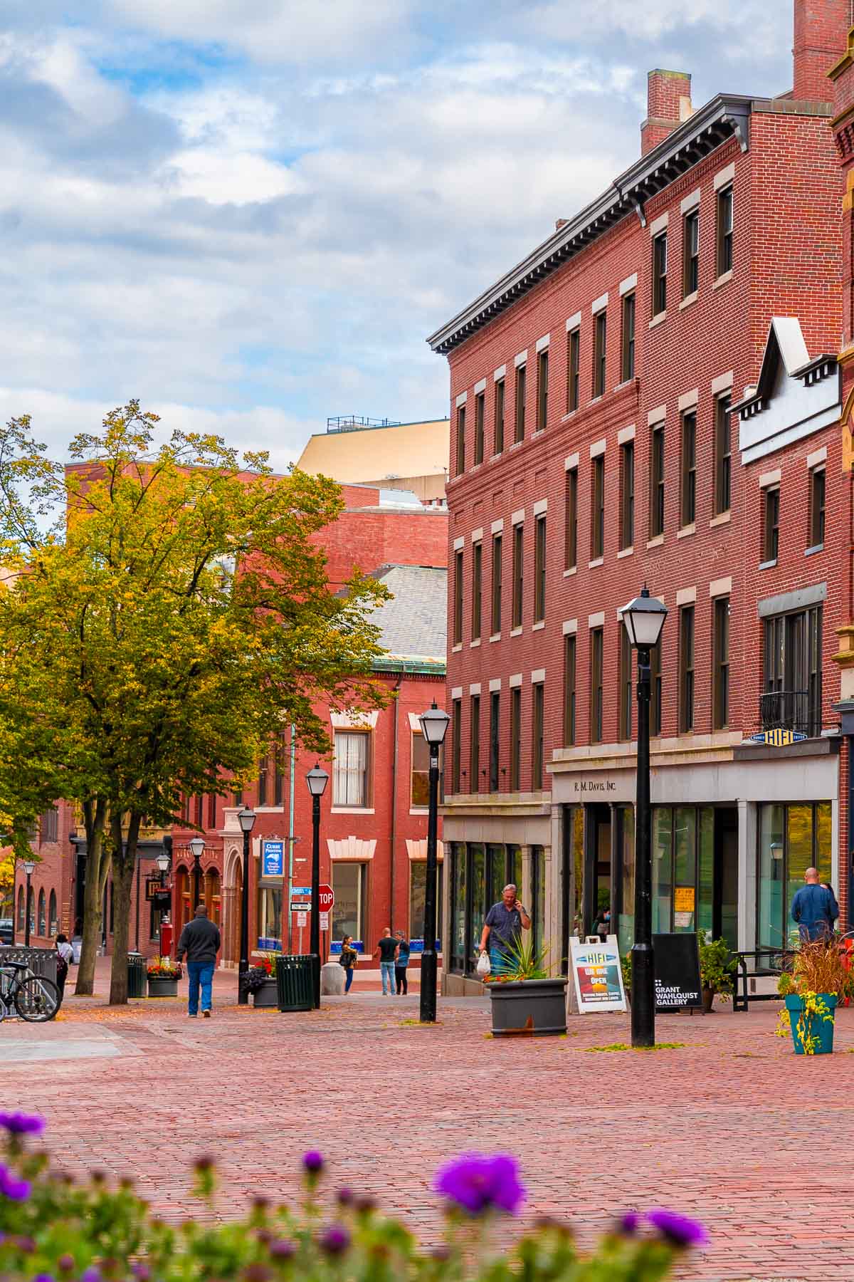 People walking along a brick street lined with red brick buildings and trees starting to turn for fall in Monument Square, Portland, Maine.