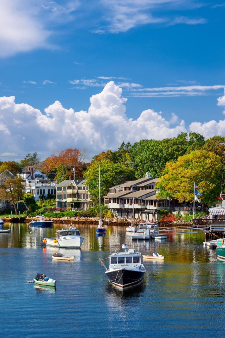 Boats floating in the harbor in Ogunquit, Maine, with colorful houses and trees lining the shoreline in the background.